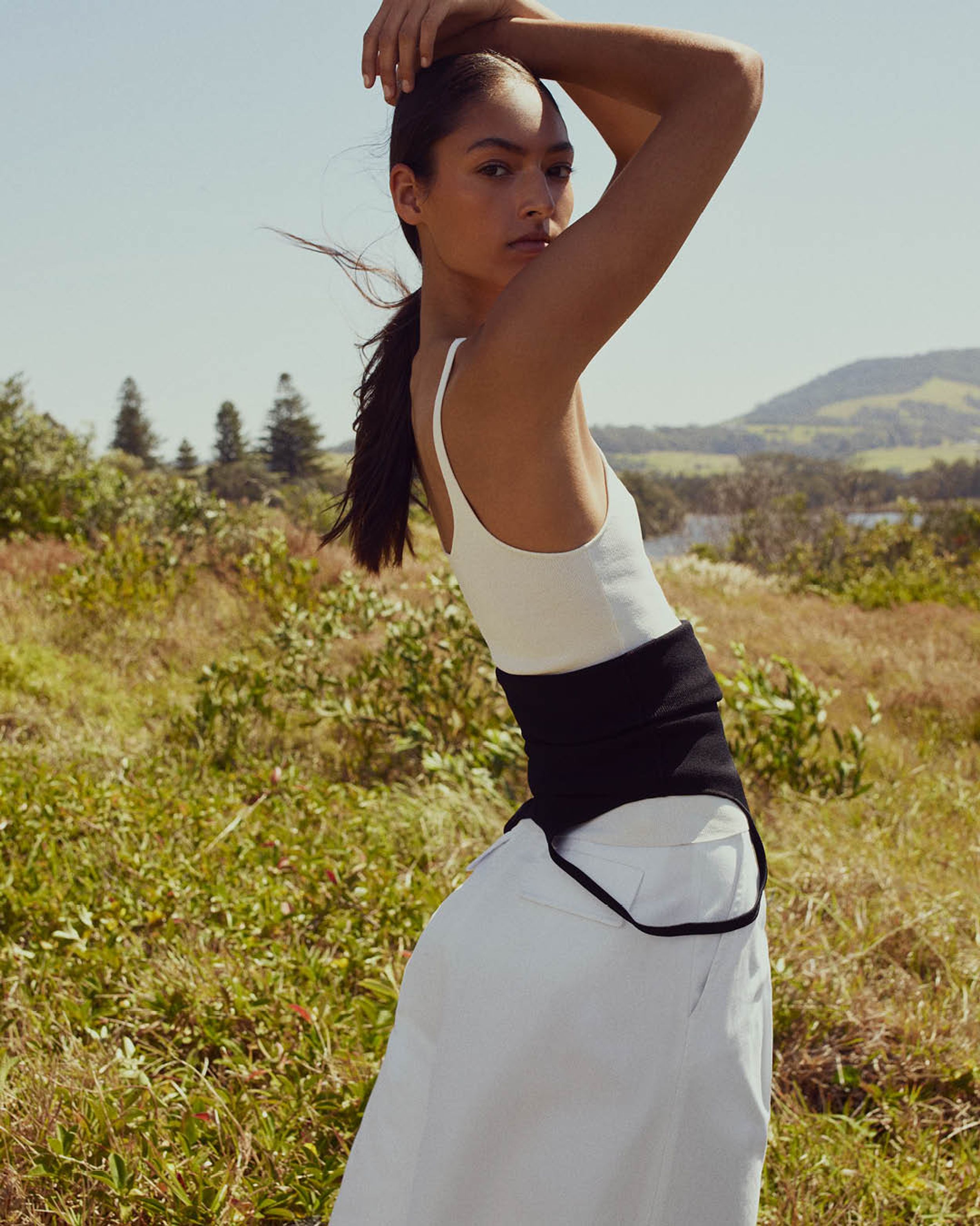 Model standing on grass with hands above her head wearing a white singlet and white wide-leg trousers