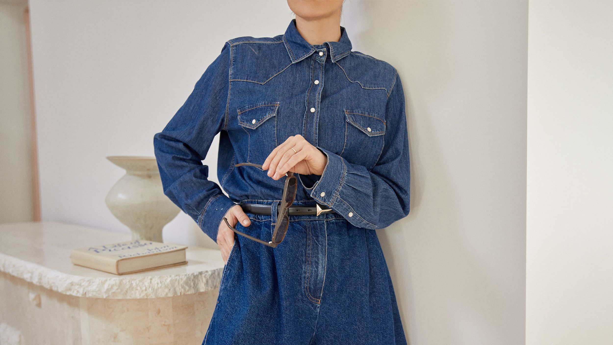 Double denim outfit of button up long sleeve shirt and matching shorts, holding sunglasses to accessorise the look