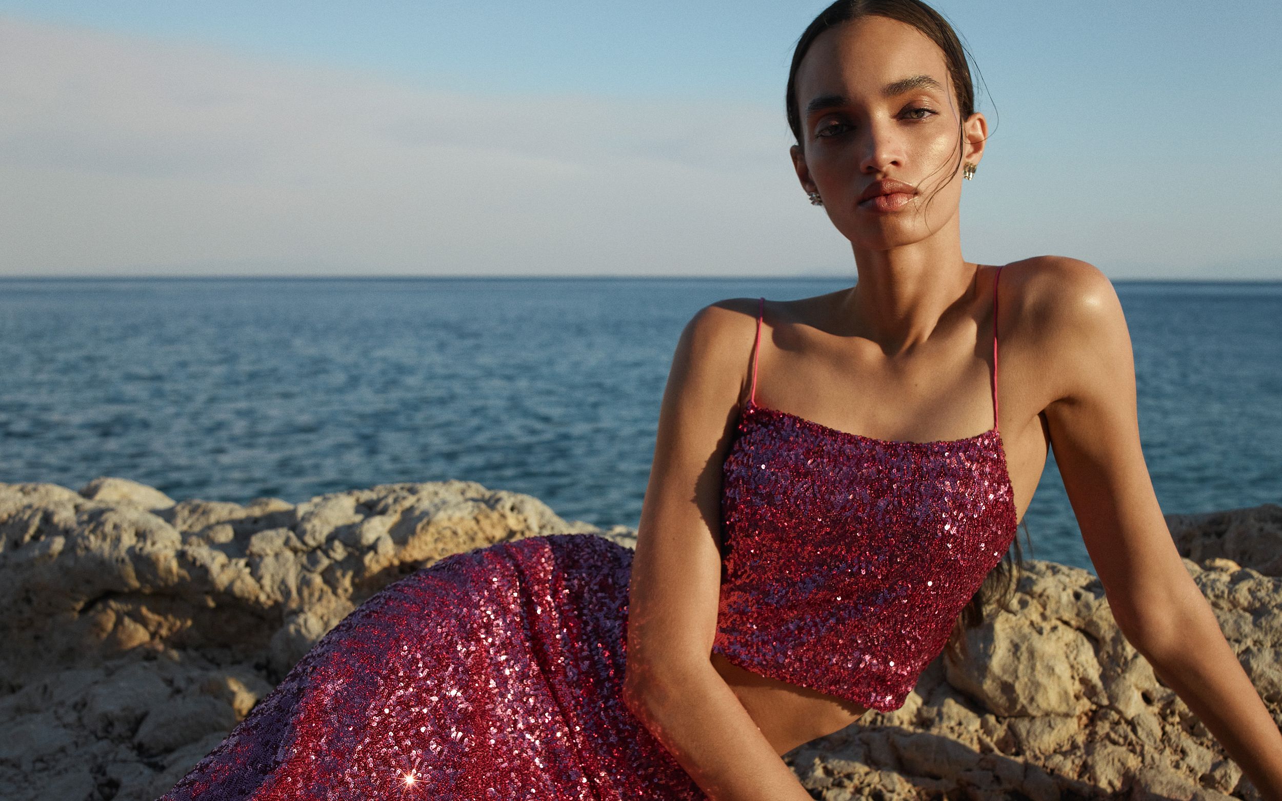 Model wearing a pink sequined singlet and skirt sitting on rocks with the ocean in the background