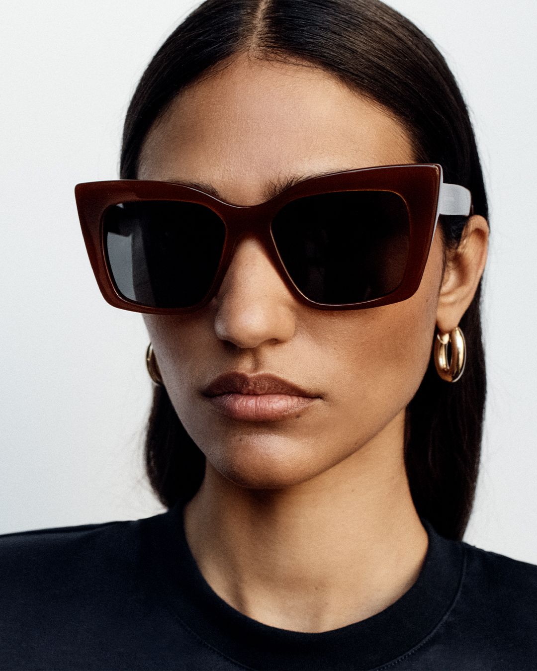 Model facing the camera wearing brown oversized sunglasses and gold hoop earrings