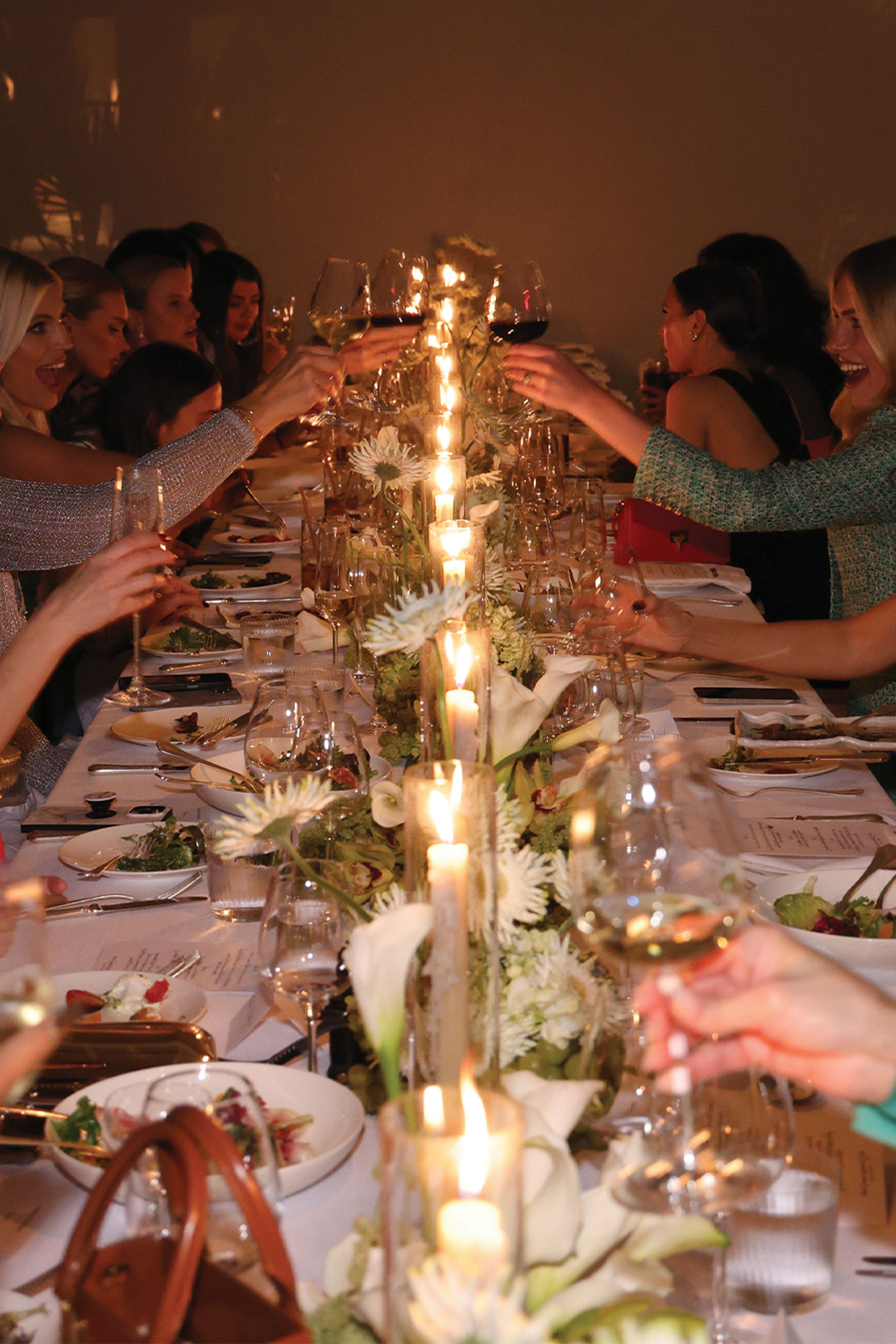 Table setting with a long row of candles and various floral arrangements with people holding wine glasses