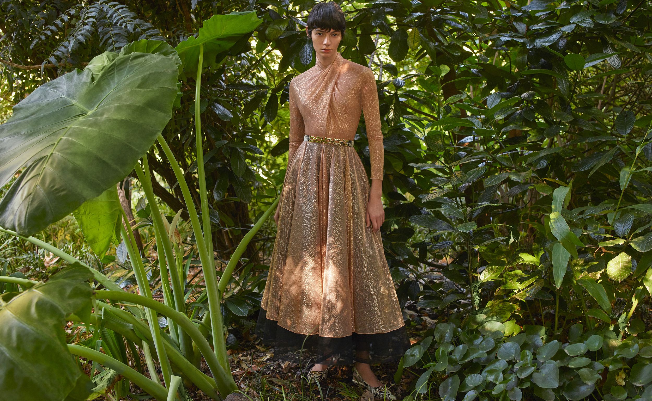 Model standing amongst dense greenery wearing a shimmering peach-coloured long-sleeved gown