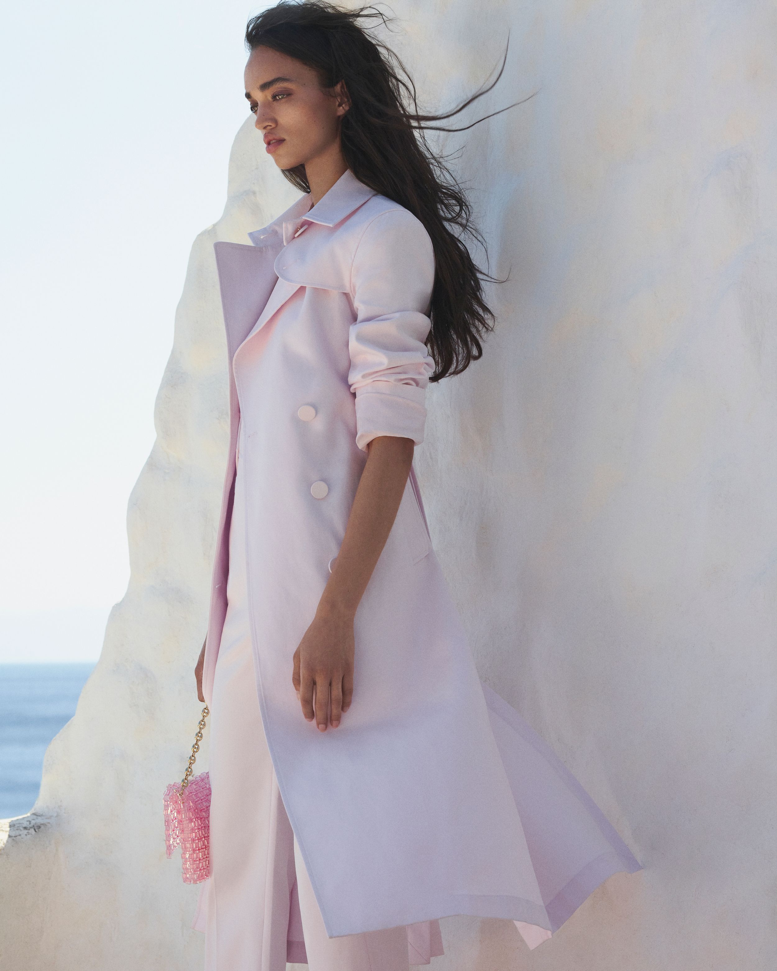 Model standing outside facing the side wearing a baby pink trench coat with the ocean in the background