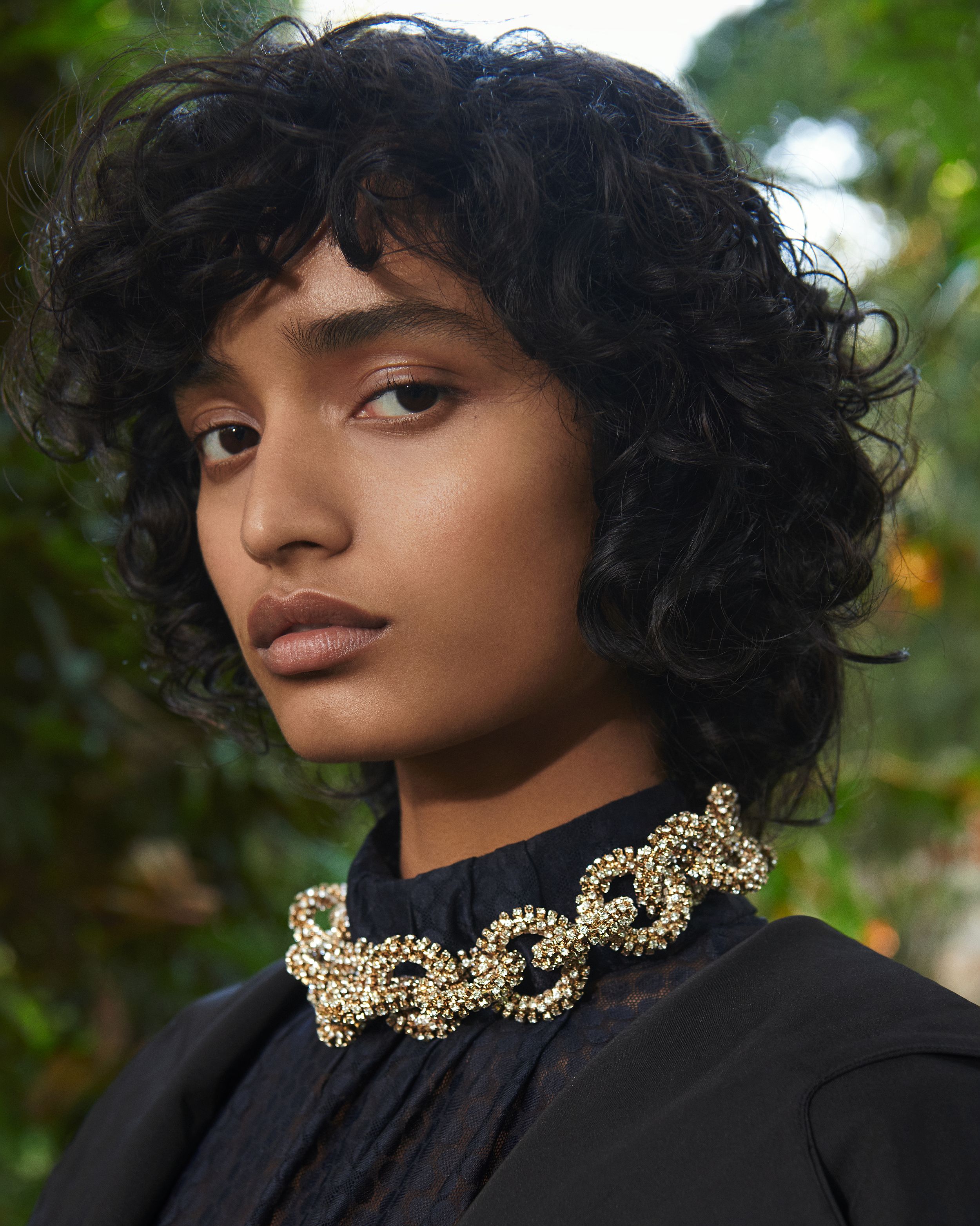 Headshot of a model with curly brown hair wearing a black high-neck blouse and chunky gold diamante chain necklace