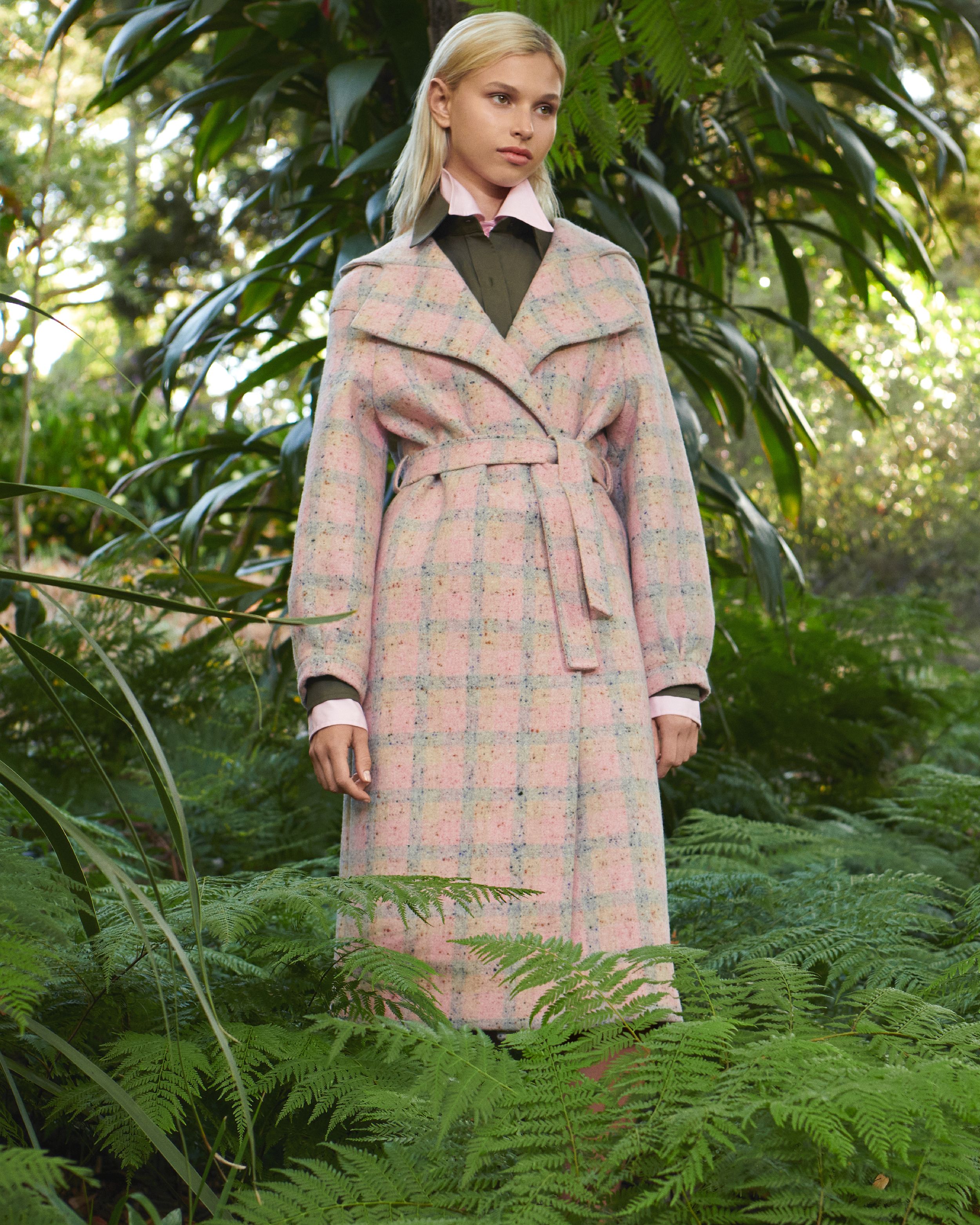 Model standing amongst greenery wearing a pink checked coat with khaki and pink shirts layered underneath