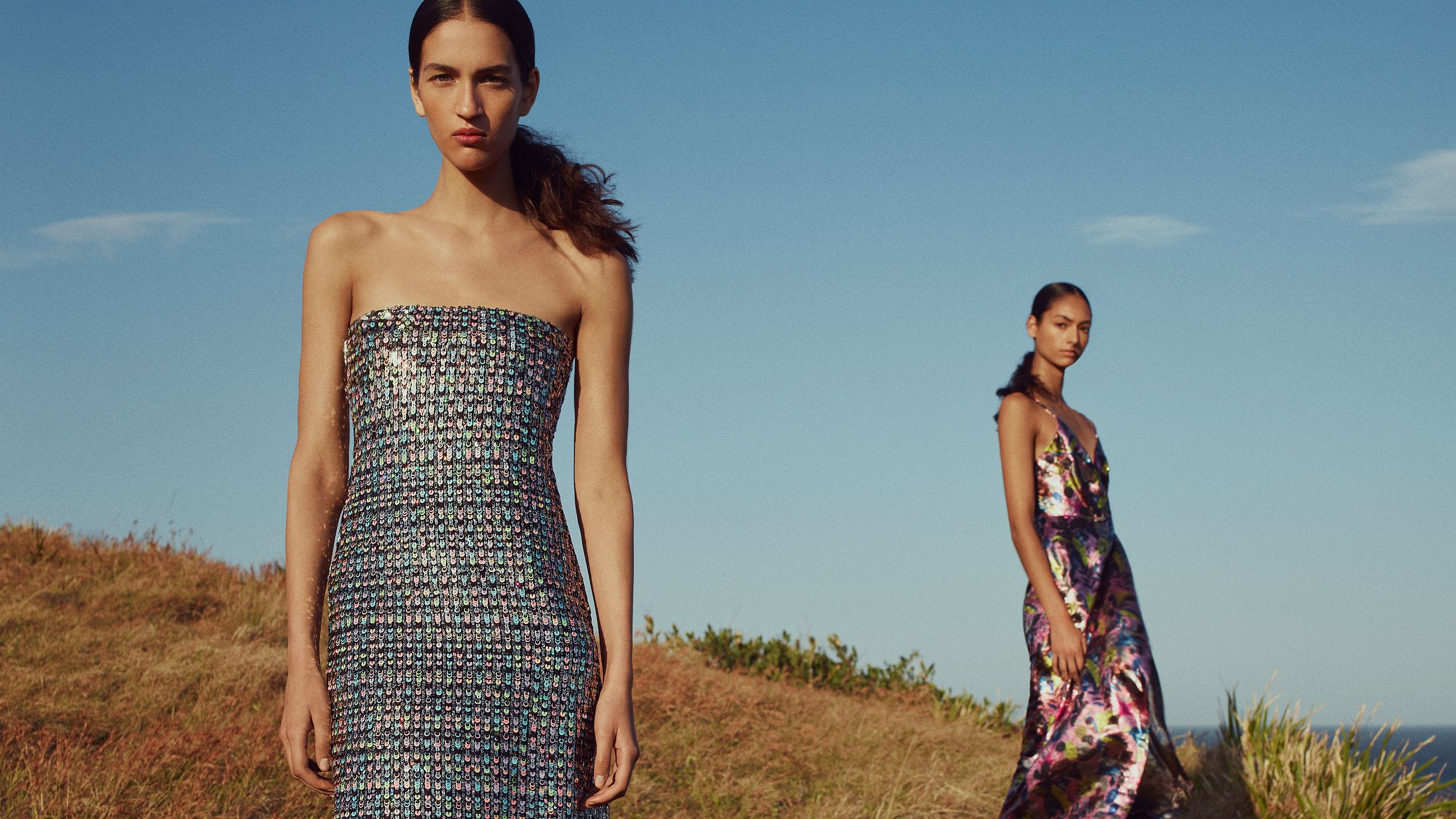 Two models standing on grass gazing at the camera wearing multicoloured shimmering evening gowns