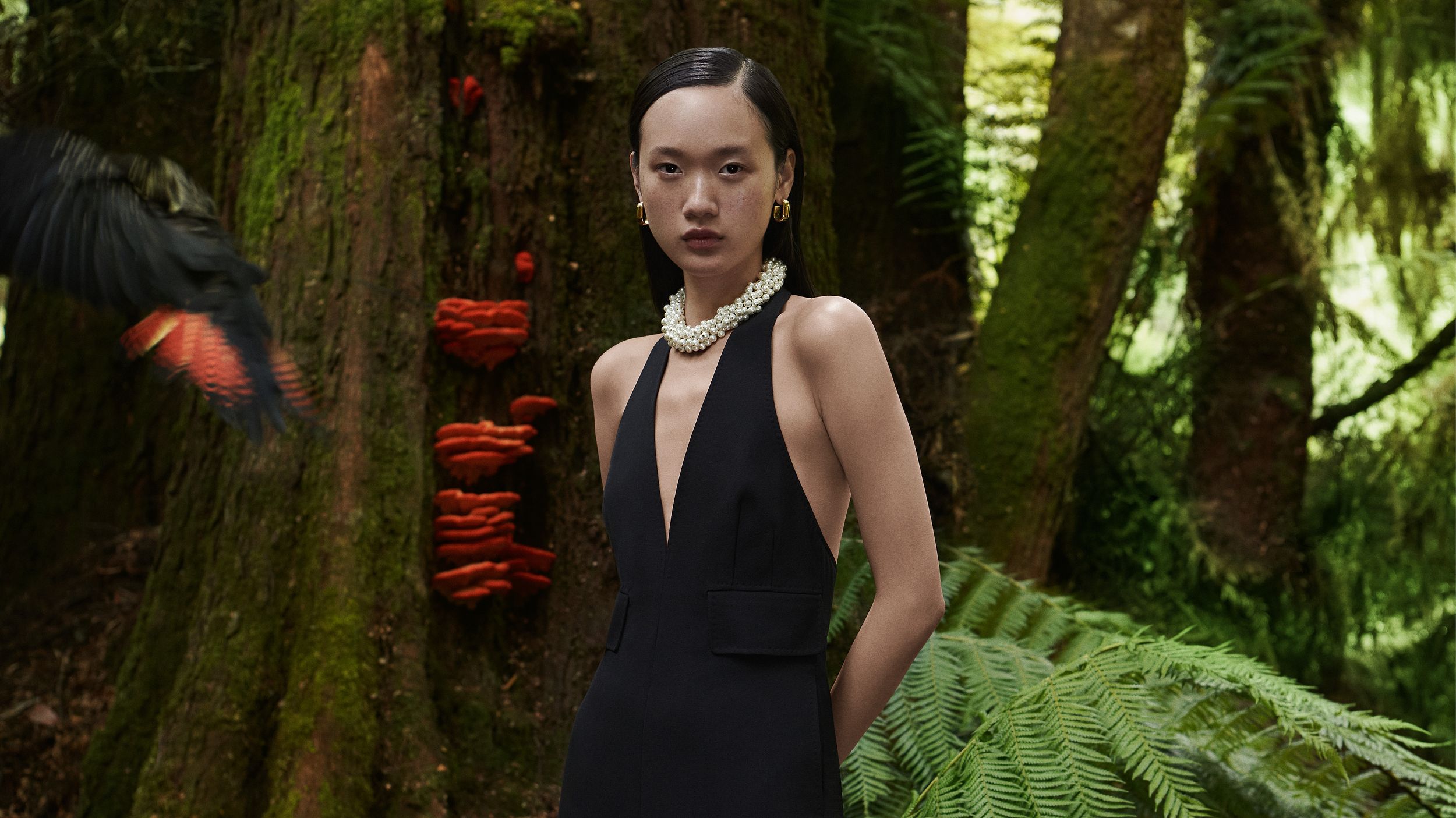 Venus He standing in the forest next to a black and orange bird wearing a black halterneck jumpsuit