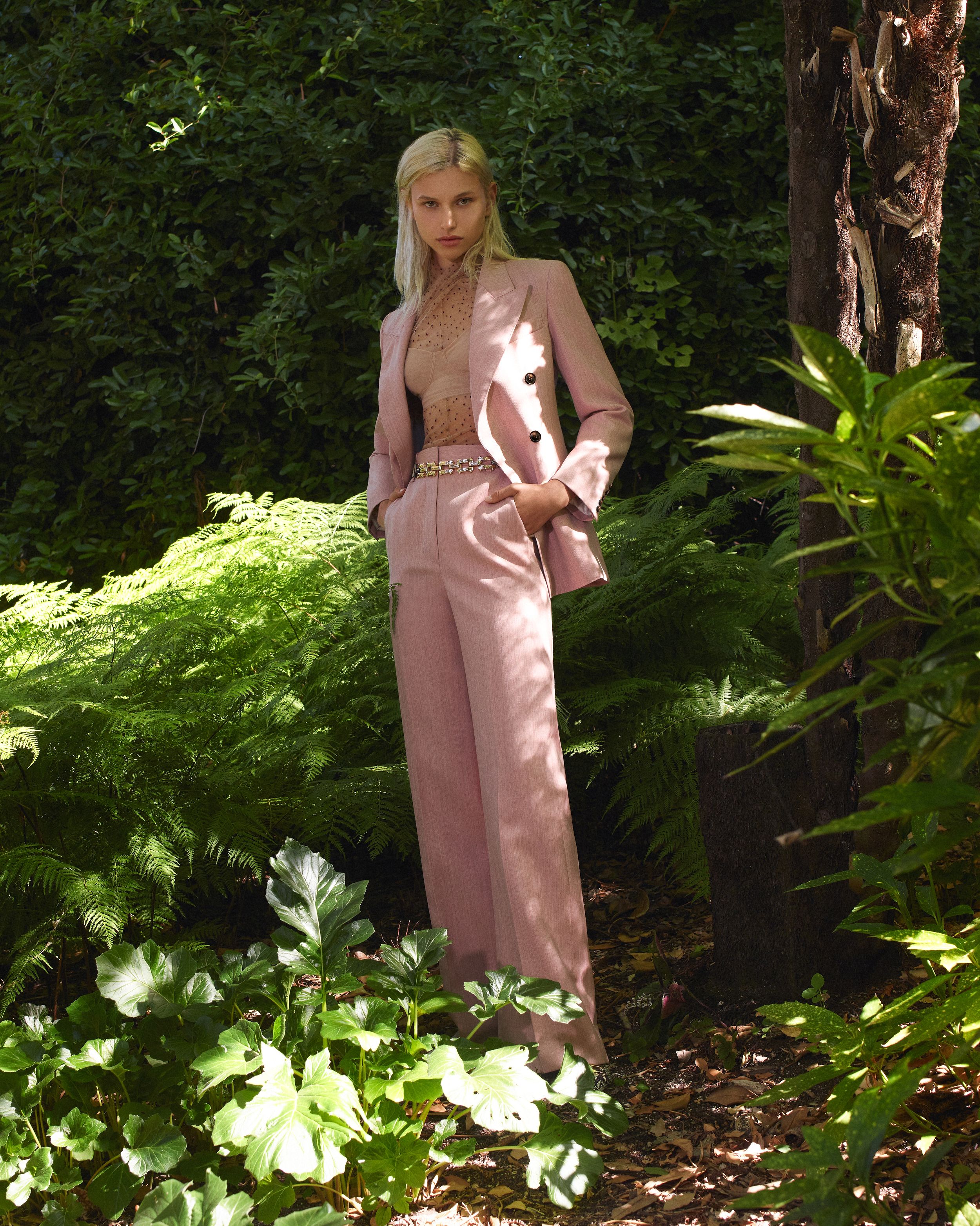 Model standing in front of dense greenery wearing a pink suit jacket and trousers