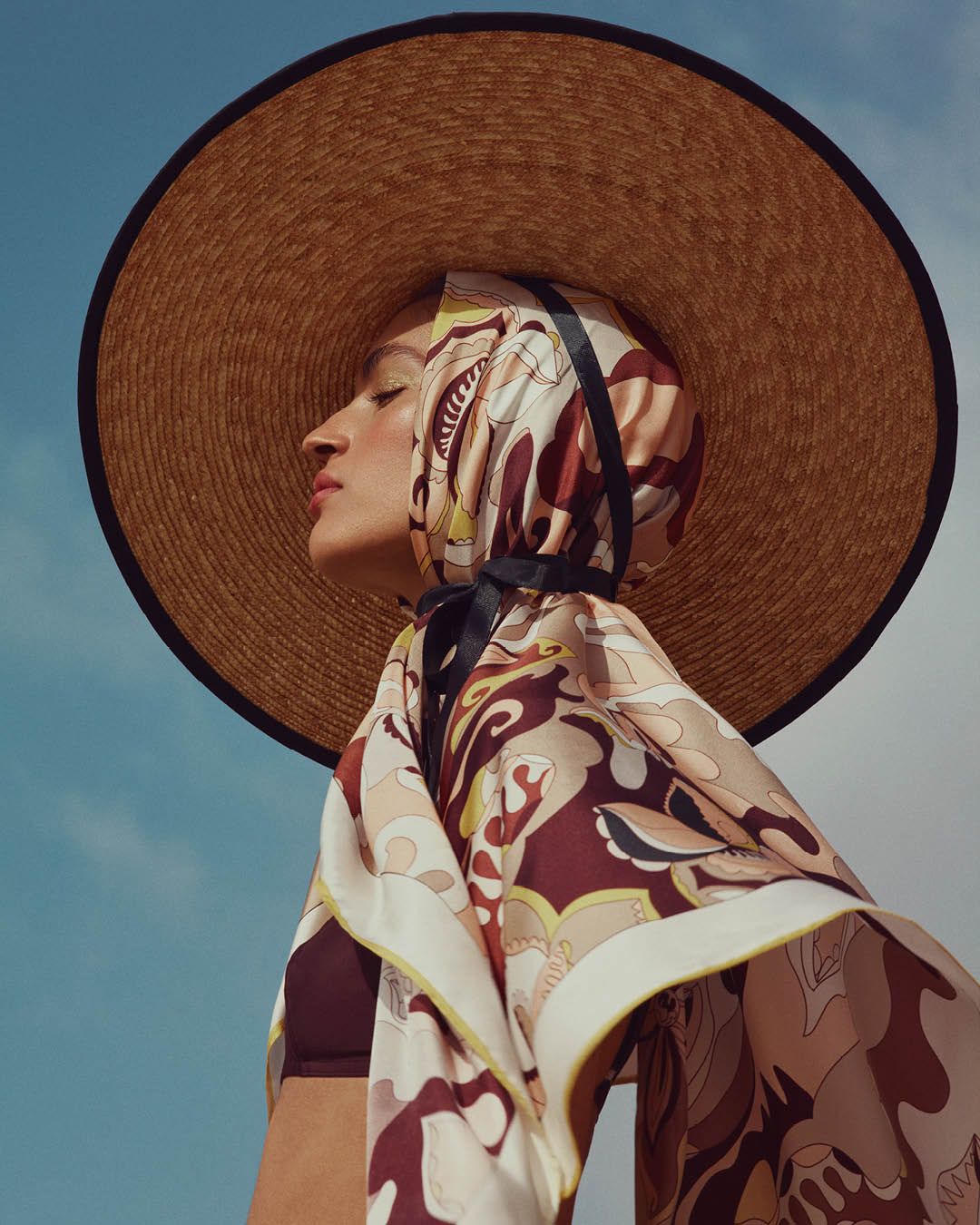 Model facing the side with eyes closed wearing straw hat with a brown, yellow and white scarf over her head and shoulders