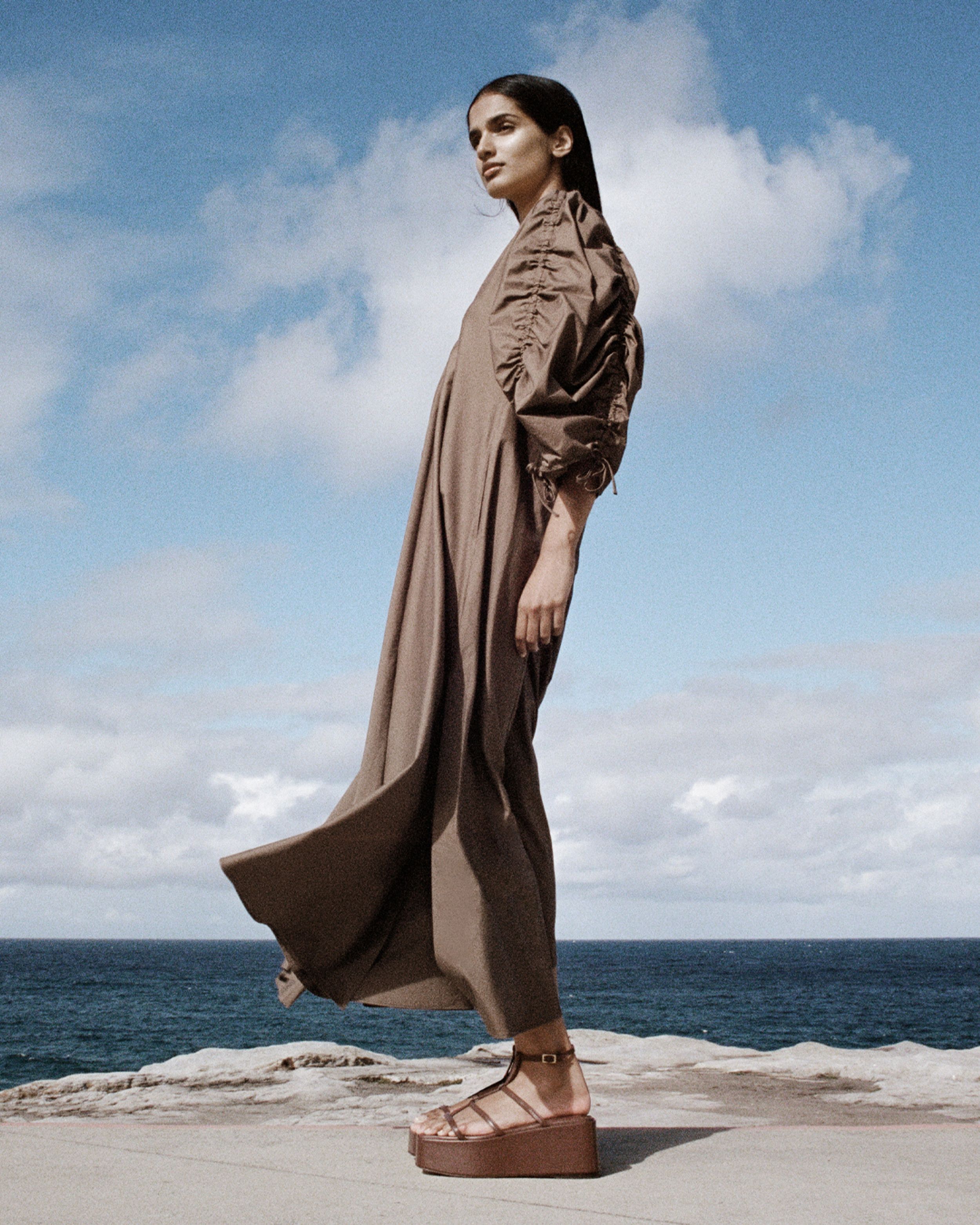 Girl standing side on, looking away from the camera, with her brown dress blowing in the wind. 