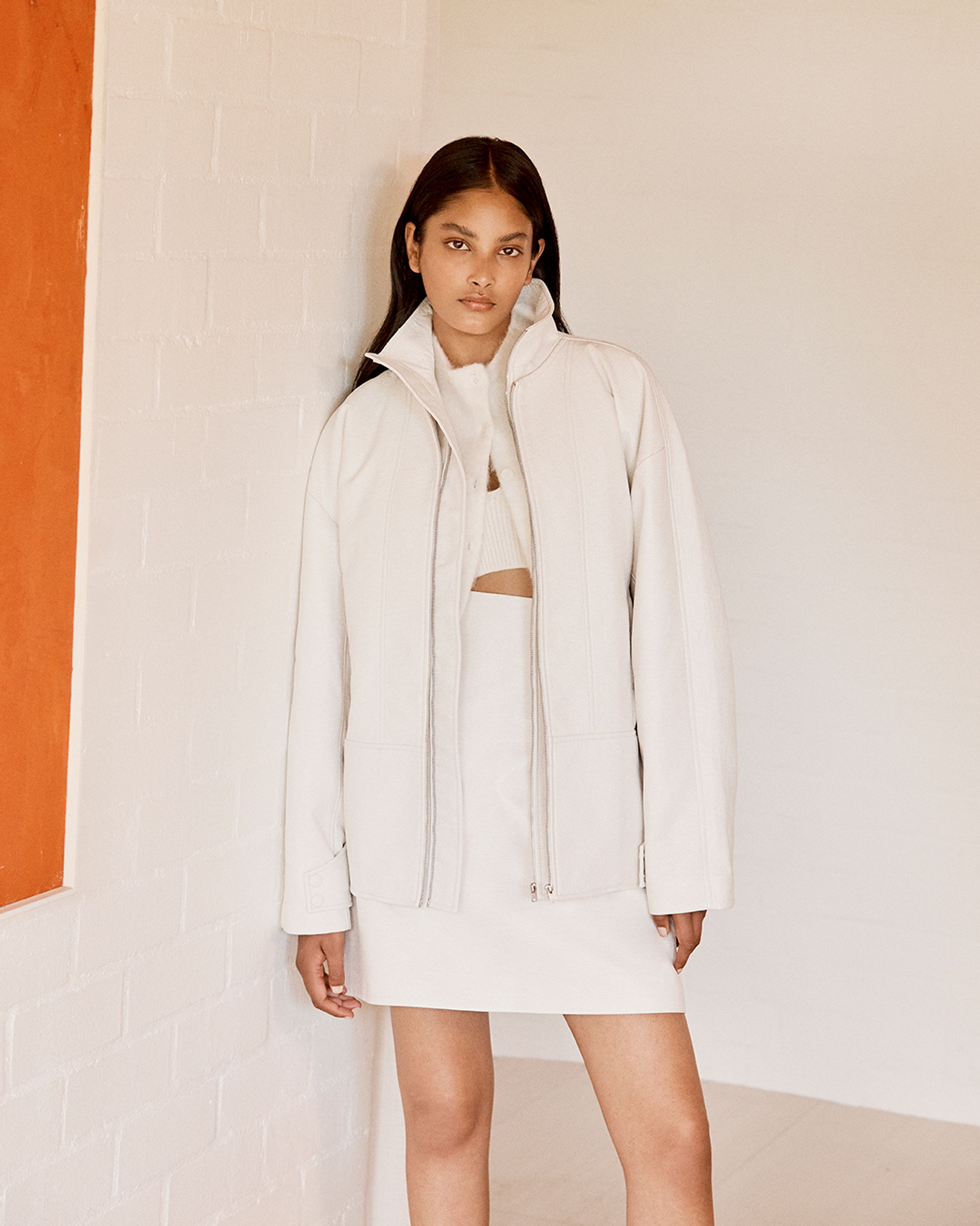 Brunette model standing wearing a white zip jacket and skirt