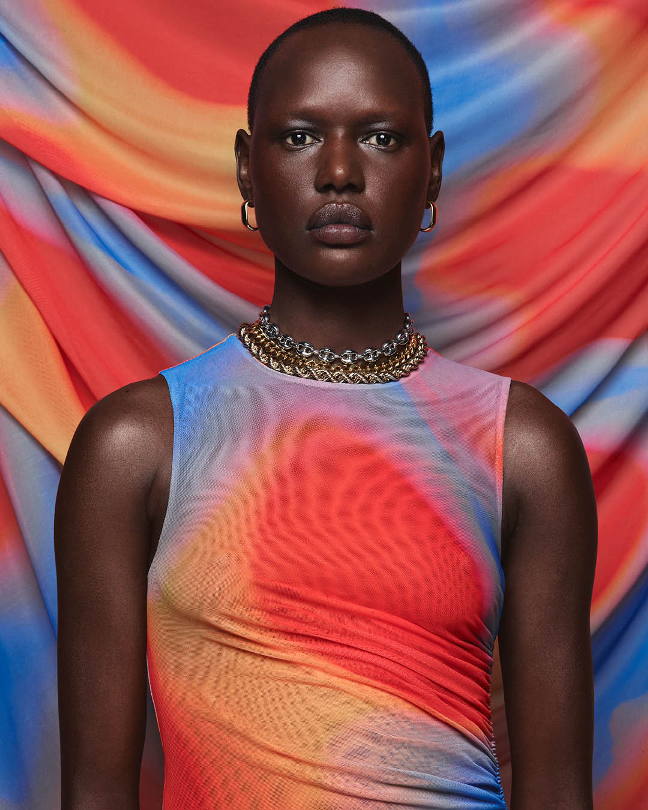 Ajak Deng wears a red, orange and blue mesh sleeveless dress standing in front of a backdrop with the same material