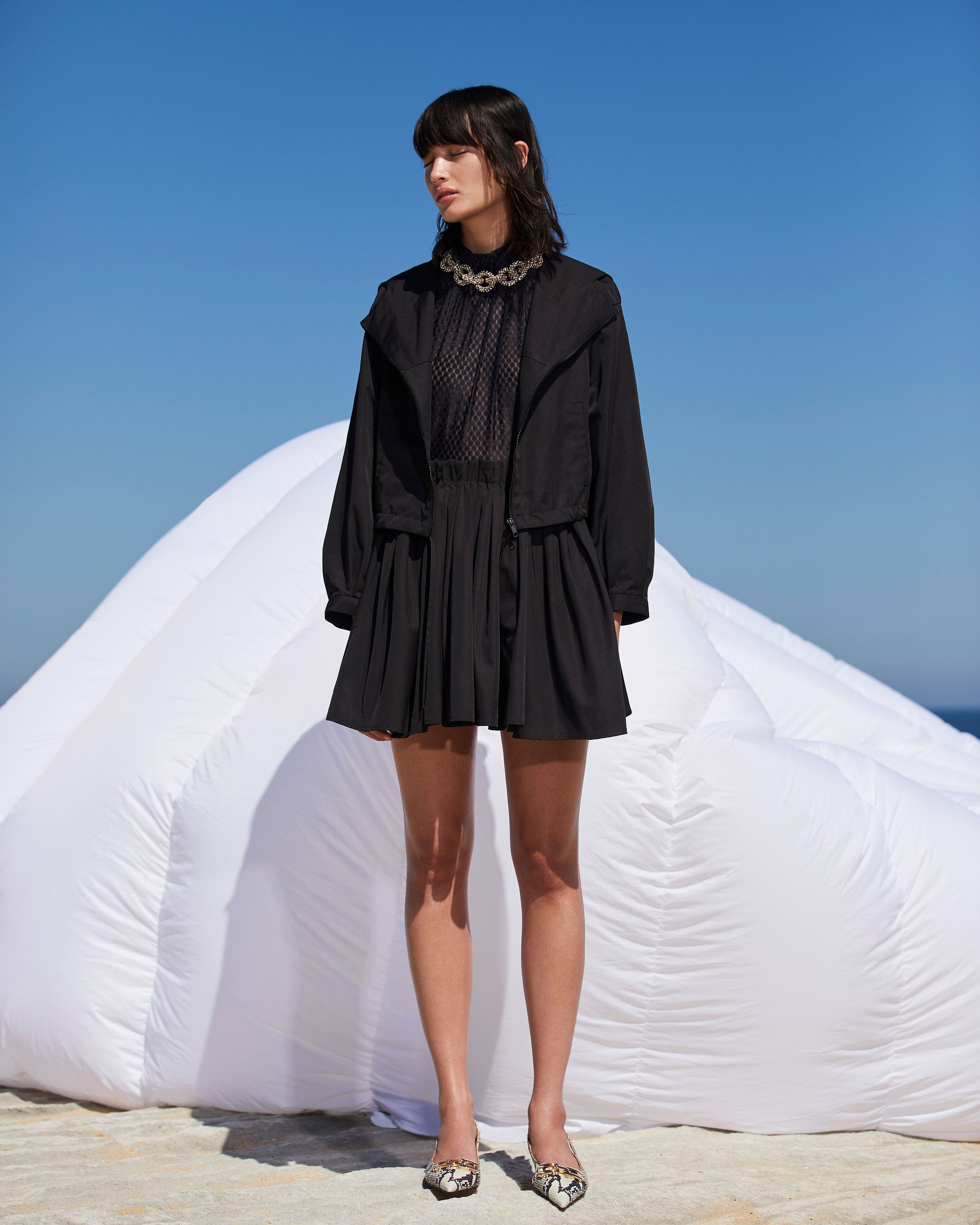 Model standing in front of a white inflatable structure wearing a black top, jacket and skirt