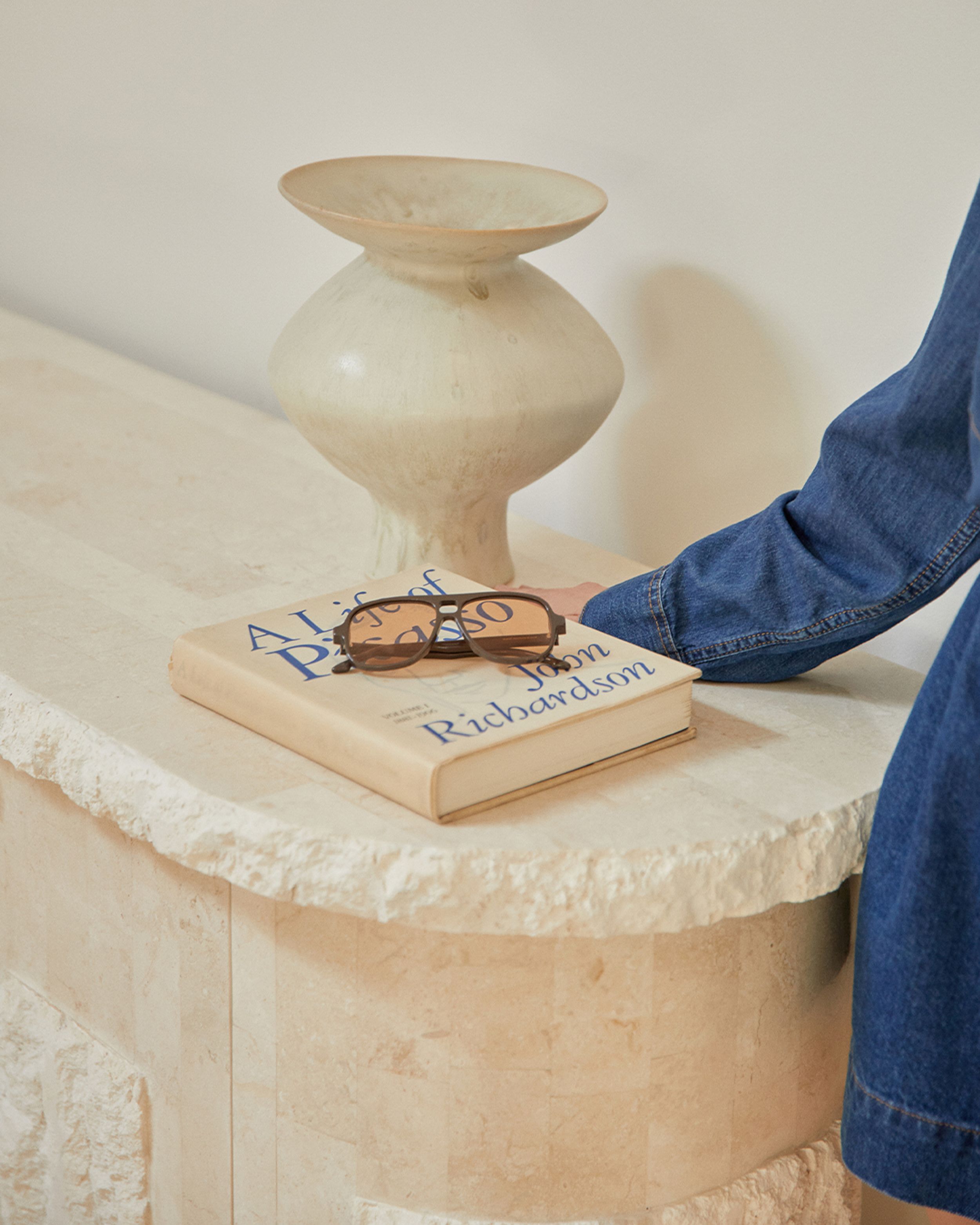 Sunglasses on top of a book with a vase beside and denim outfit.