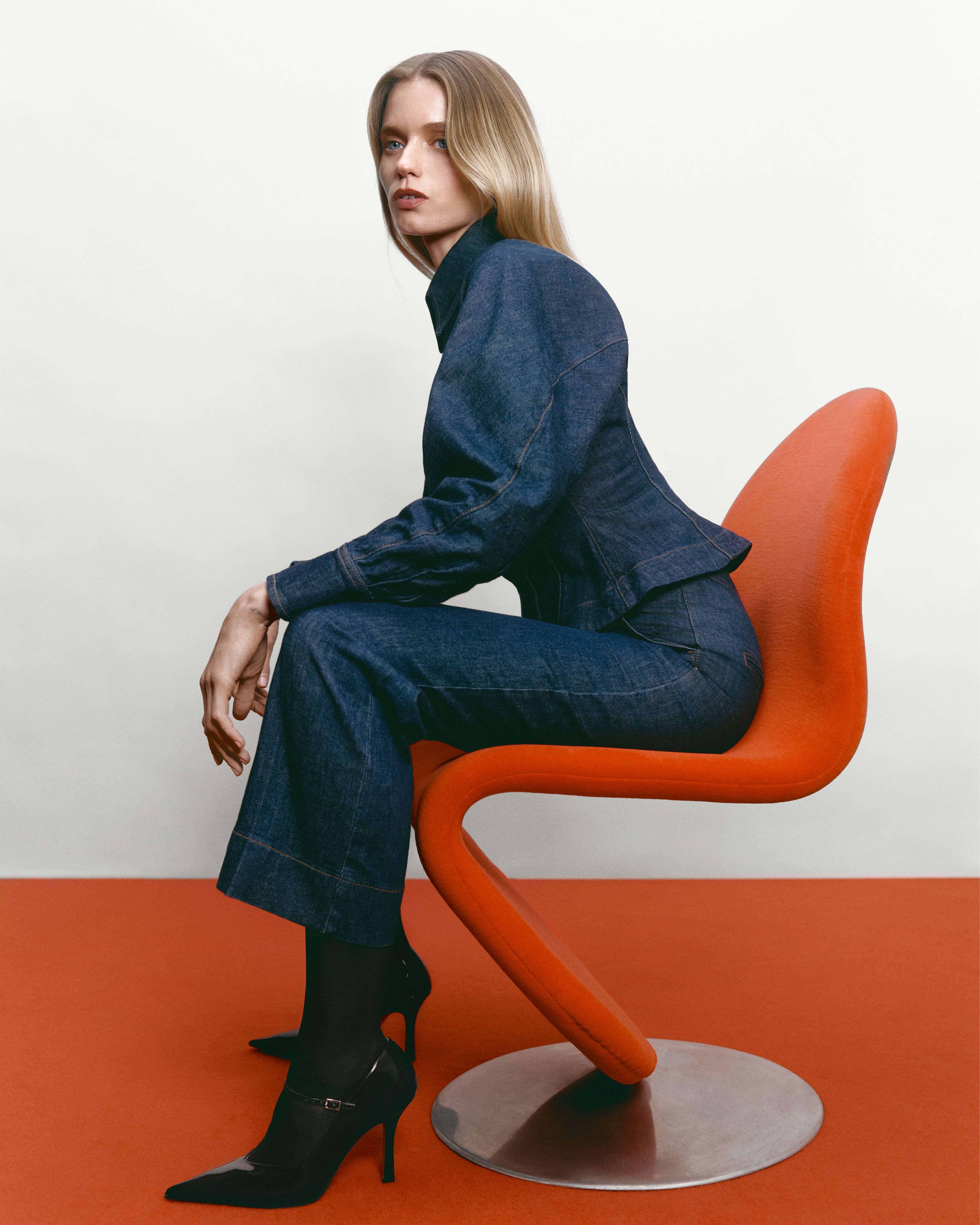 Abbey Lee sitting in a red chair facing the side wearing an indigo denim jacket and jeans
