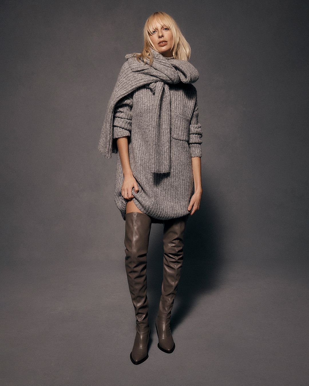 Blonde model standing wearing a grey melange knitted sweater and taupe over-the-knee boots
