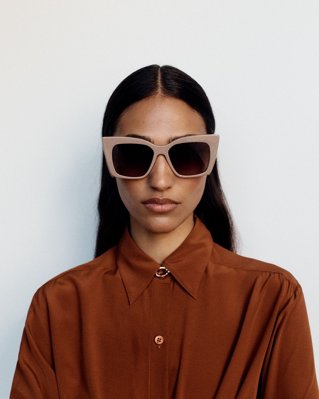 Model facing the camera wearing taupe oversized sunglasses and a rust button up silk skirt