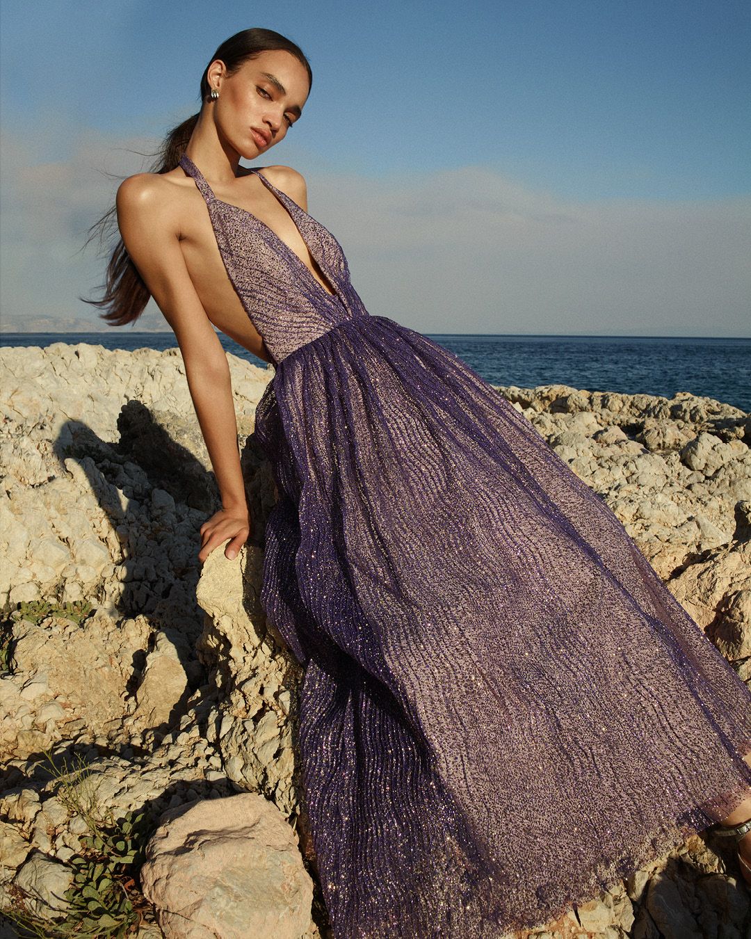 Model wearing a purple sparkly tulle gown leaning against rocks with the ocean in the background