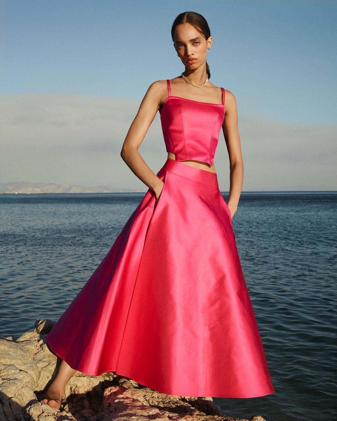 Model standing in front of the ocean wearing a matching fuschia satin bustier and high-waisted skirt