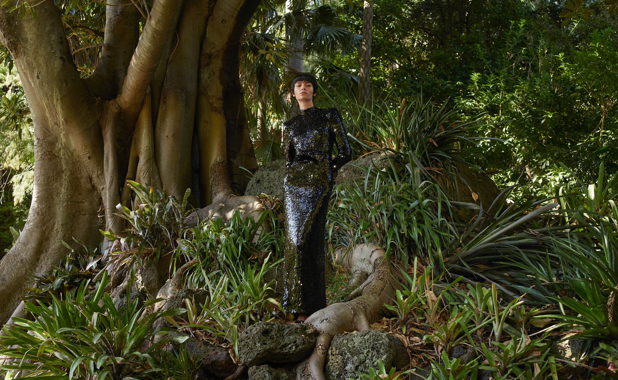 Model standing amongst dense greenery in front of a fig tree wearing a black sequined gown