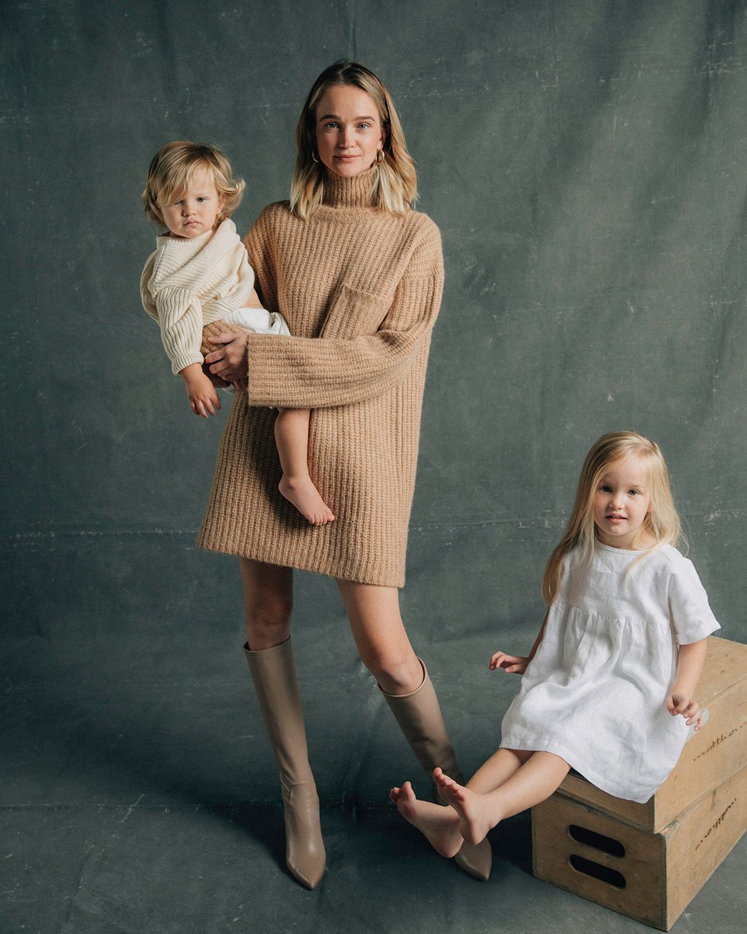 Rosie Tupper standing holding her son wearing a beige knit dress with her daughter sitting