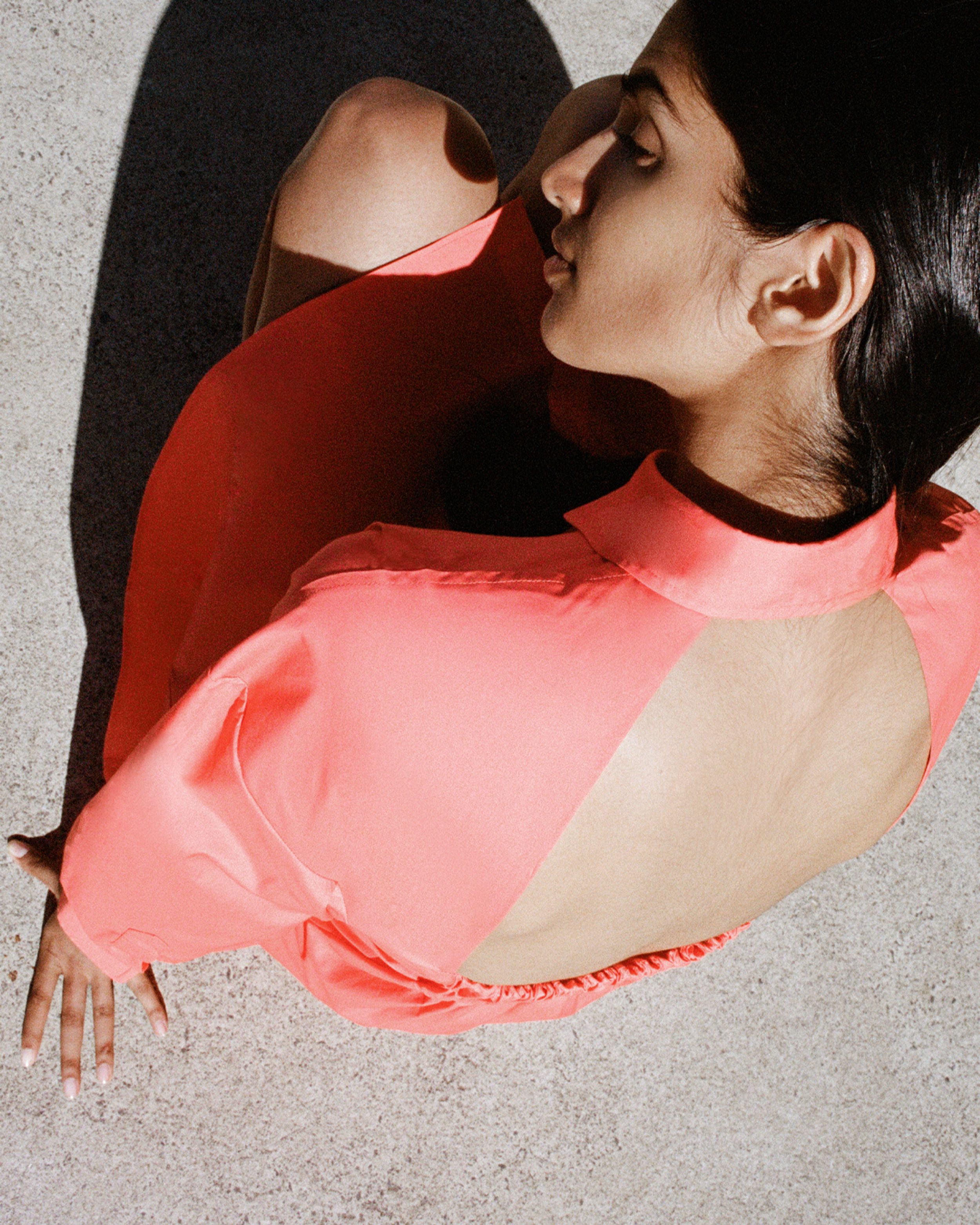 Girl crouching on the ground with head on knees looking away wearing coral dress with back cut out. 