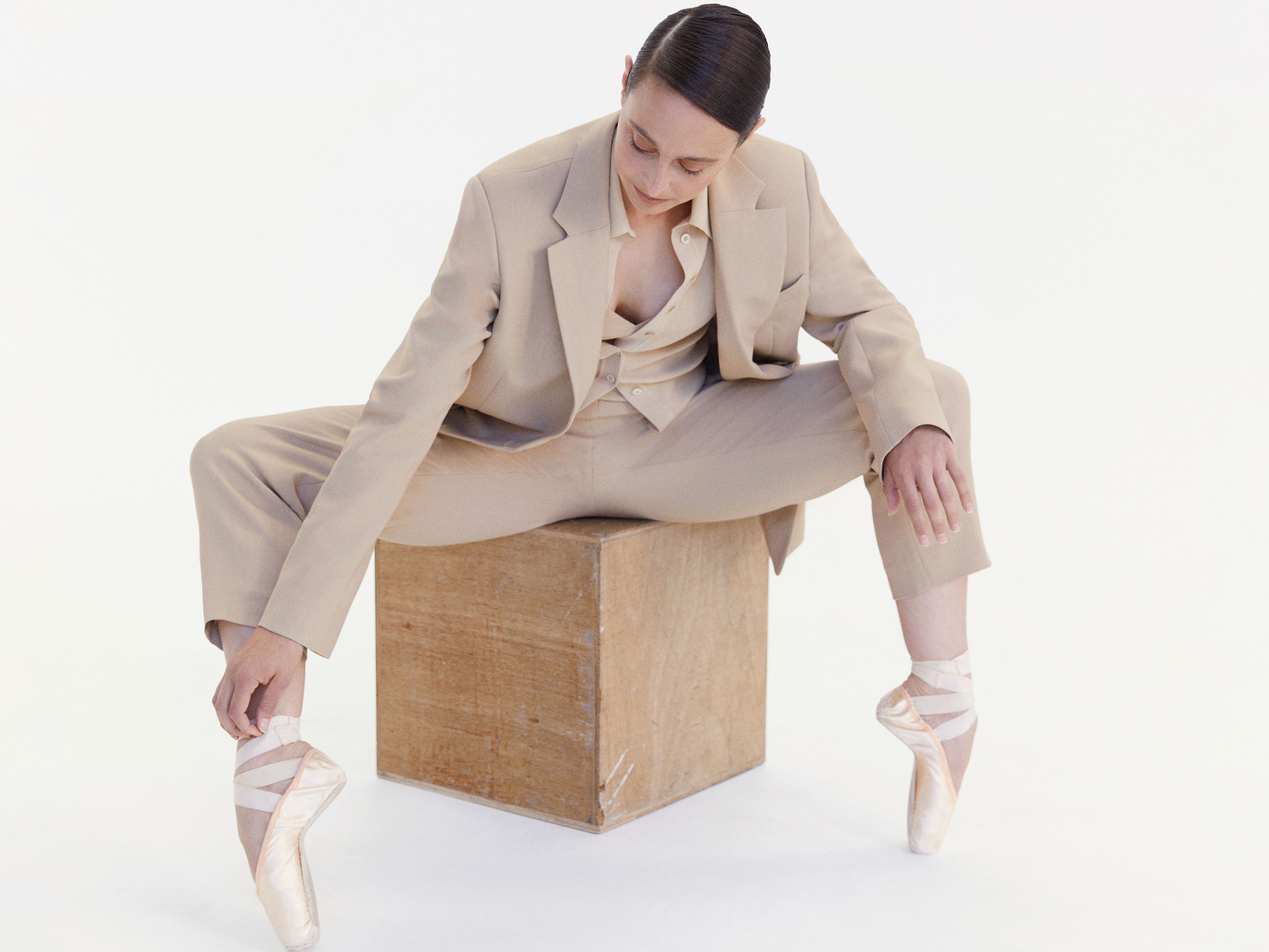 Dimity Azoury sitting on a beige cube stool wearing a beige suit and pointe ballet shoes