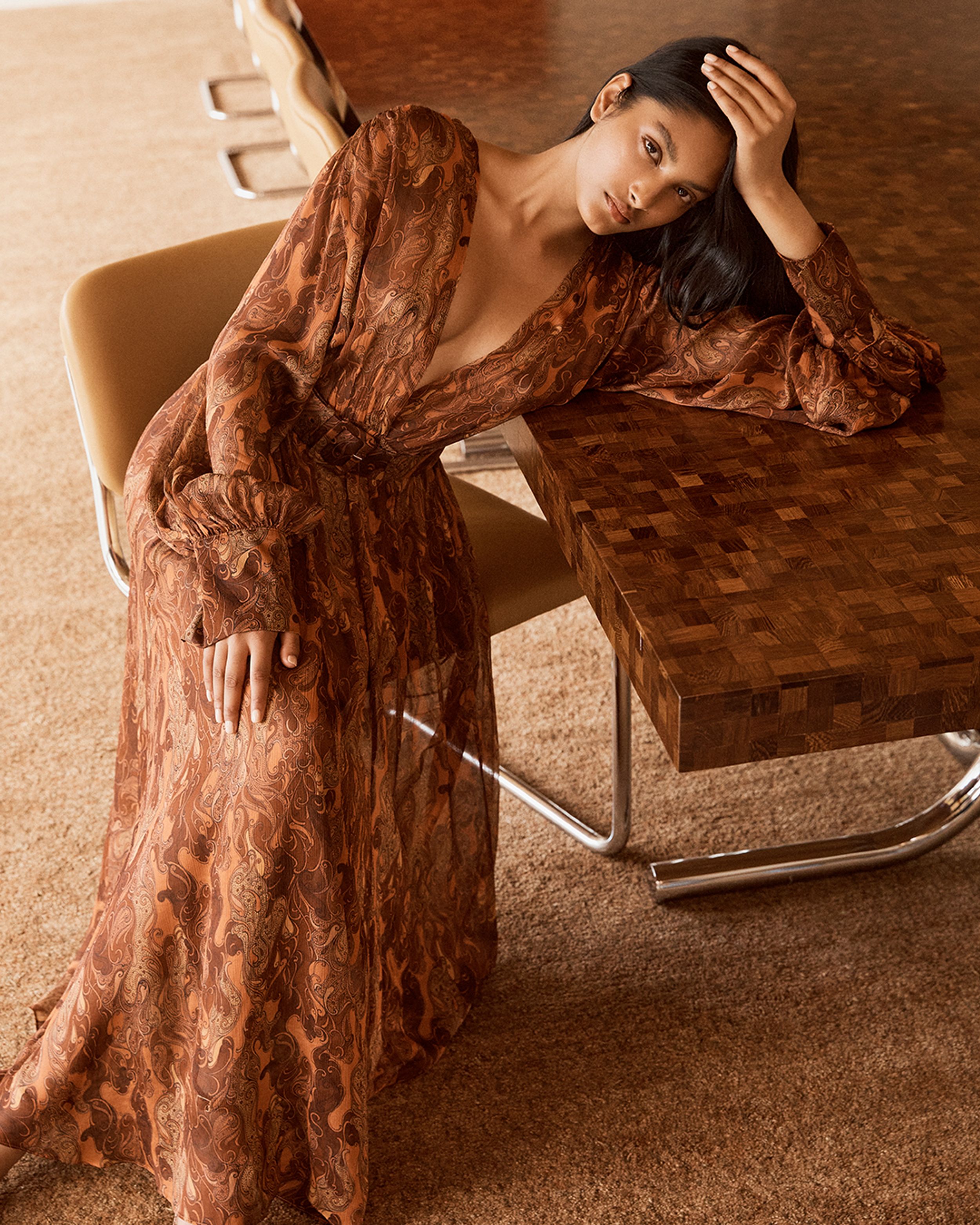 Brunette model seated leaning on a dining table wearing a brown print dress