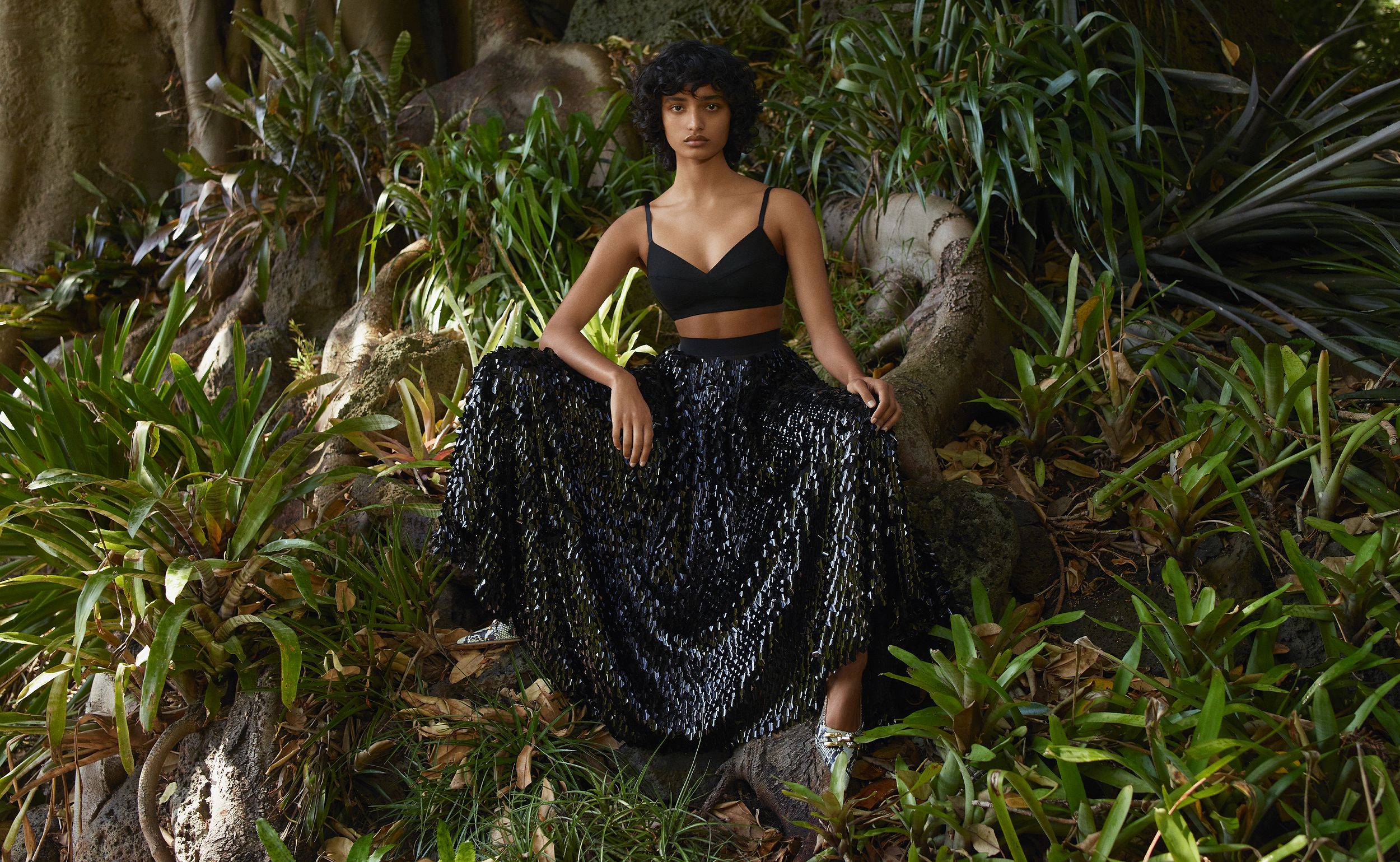 Model sitting amongst greenery wearing a black bralette and black sequined floor-length skirt
