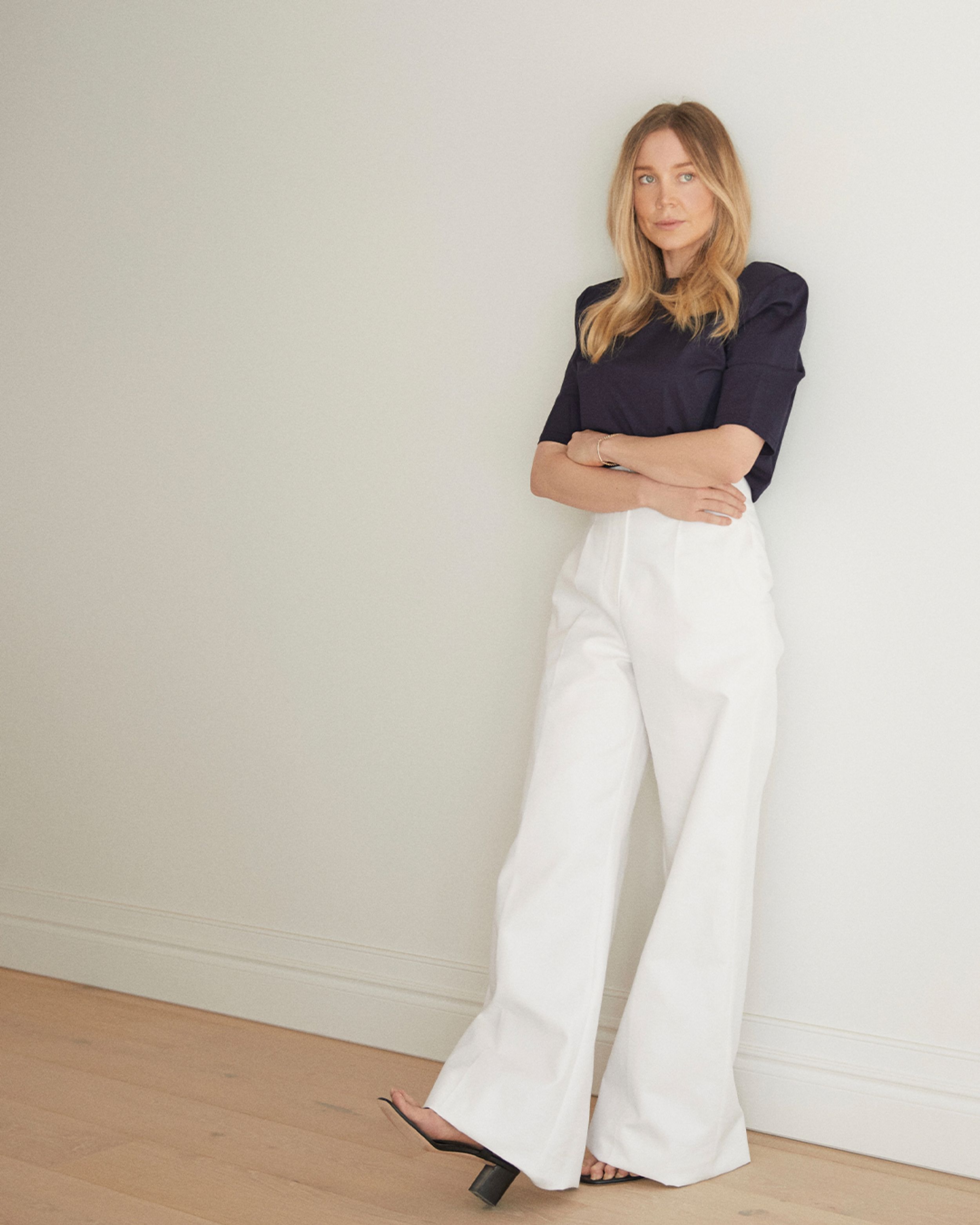 Hayley Bonham with long blonde hair leaning on a wall and crossing her arms wearing a dark short sleeve t-shirt and wide long white trousers