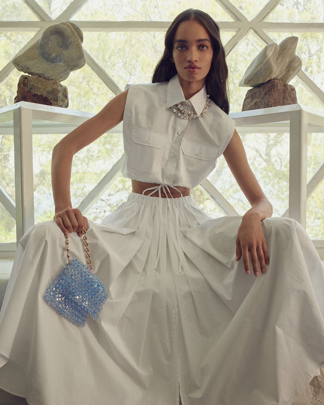 Model sitting wearing a coordinating cropped white shirt and long skirt, holding a baby blue beaded bag