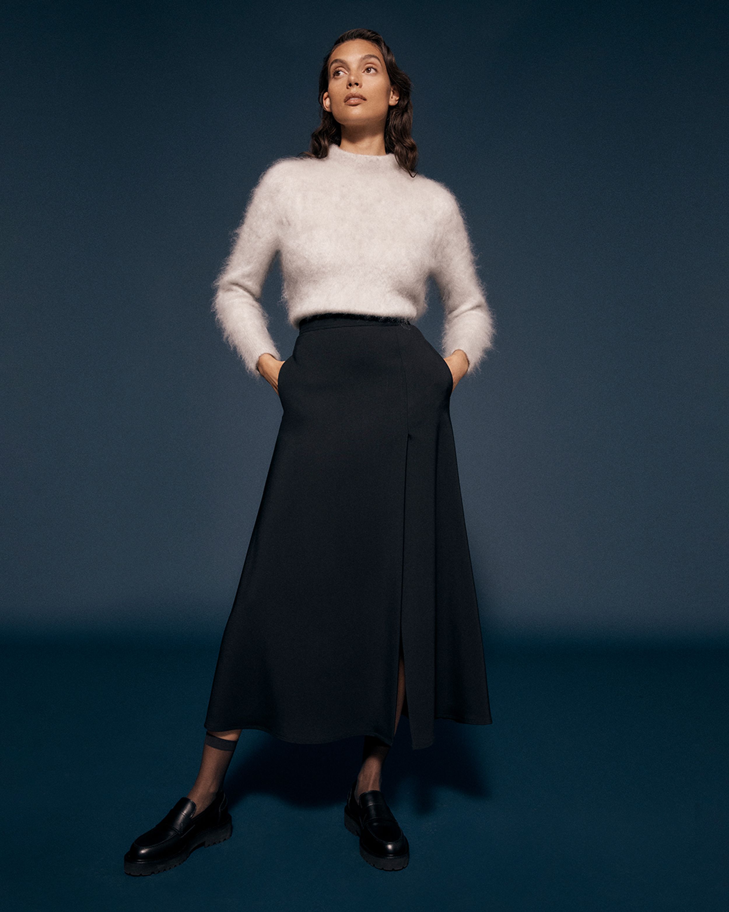 Brunette model standing with hands in pockets wearing a white mohair sweater and black skirt