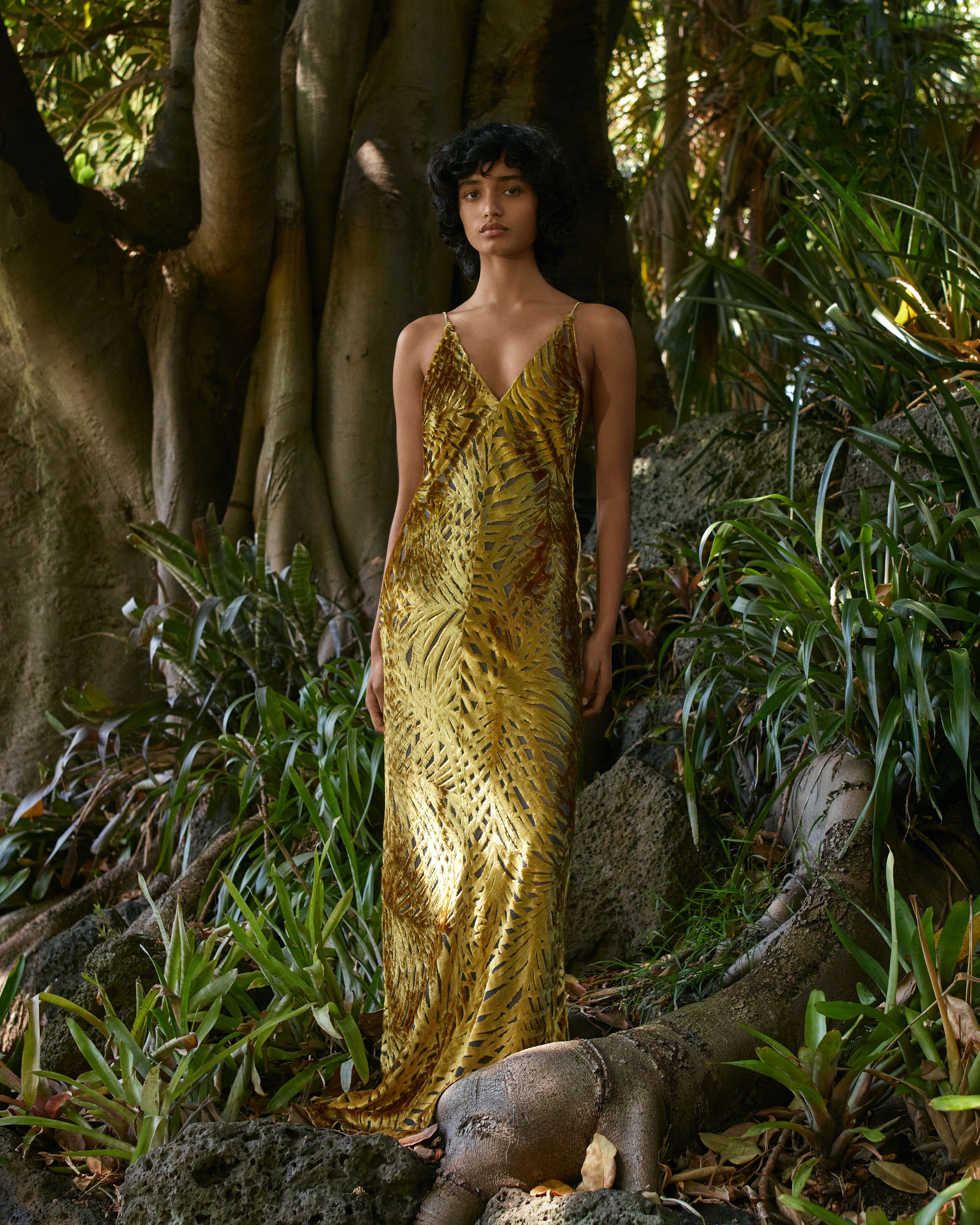 Model standing in front of a fig tree wearing a gold floor-length satin slip dress