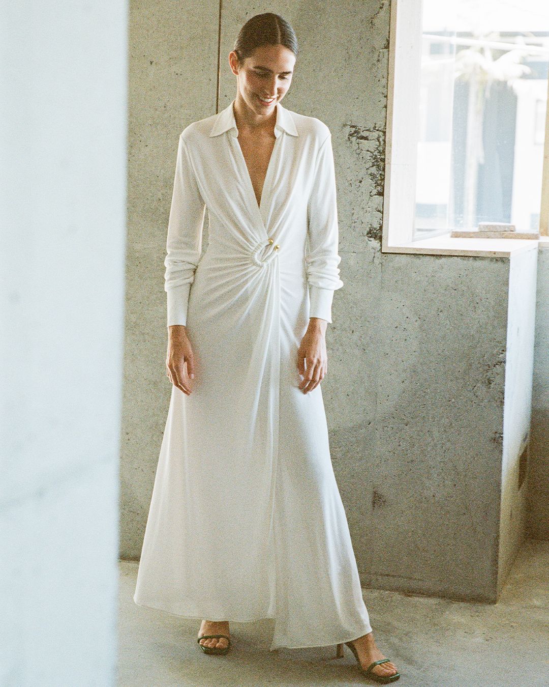 Bianca Marchi looking down standing in front of a concrete wall wearing a white gown