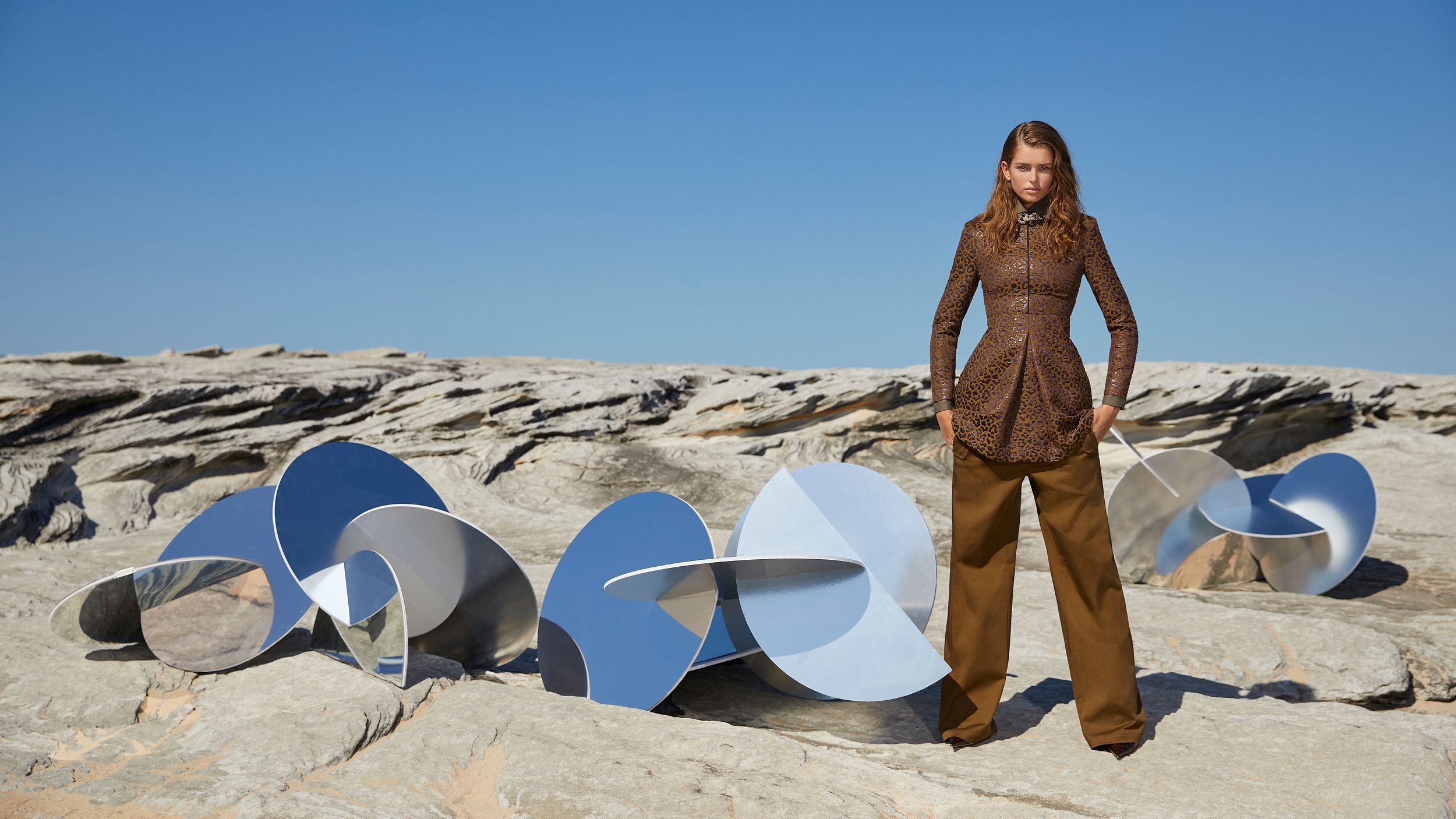 Model standing amongst mirrored sculptures outdoors wearing a brown ocelot-print dress and trousers