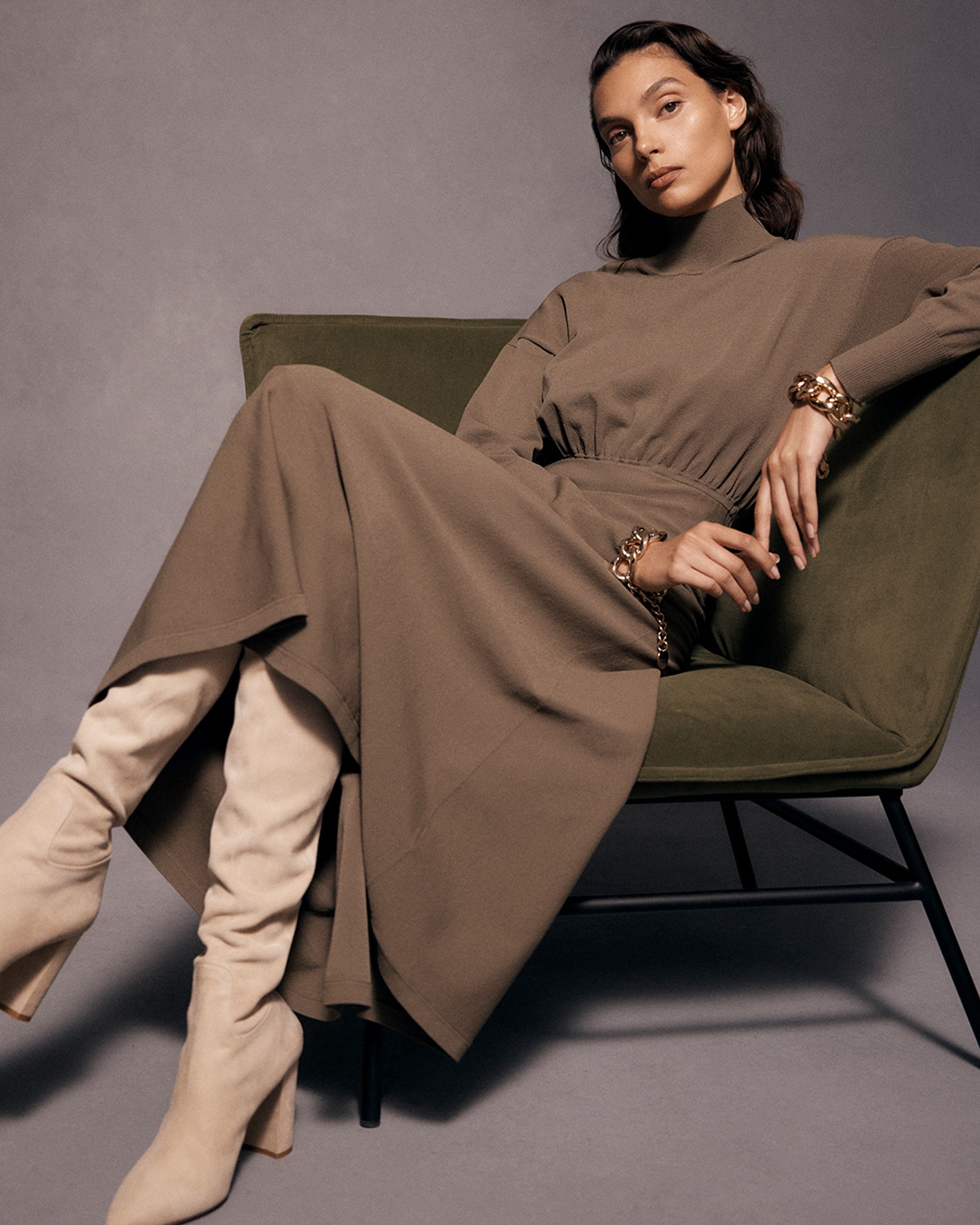 Brunette model sitting with legs crossed in an armchair wearing a brown dress