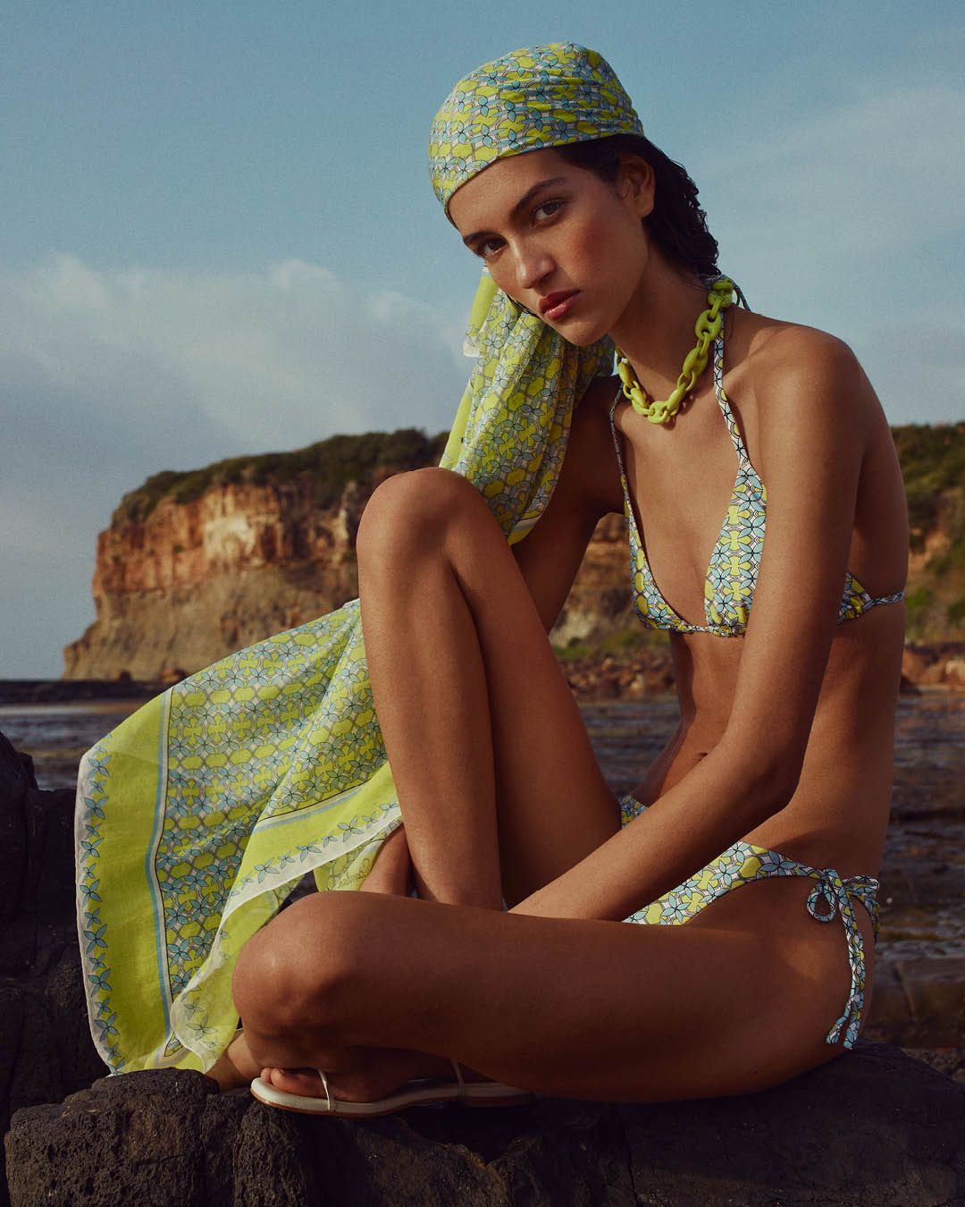 Model wearing a green and blue bikini and scarf sitting on rocks with the ocean in the background