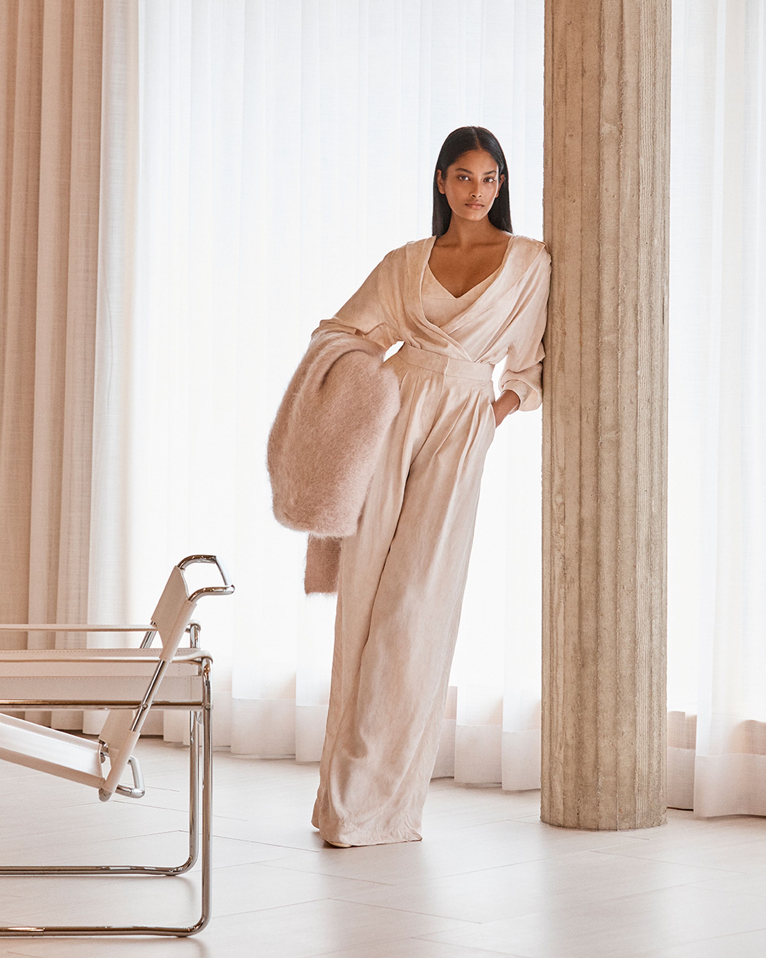 Brunette model leaning on a pillar wearing a cream-coloured shirt and trousers, holding a mohair scarf