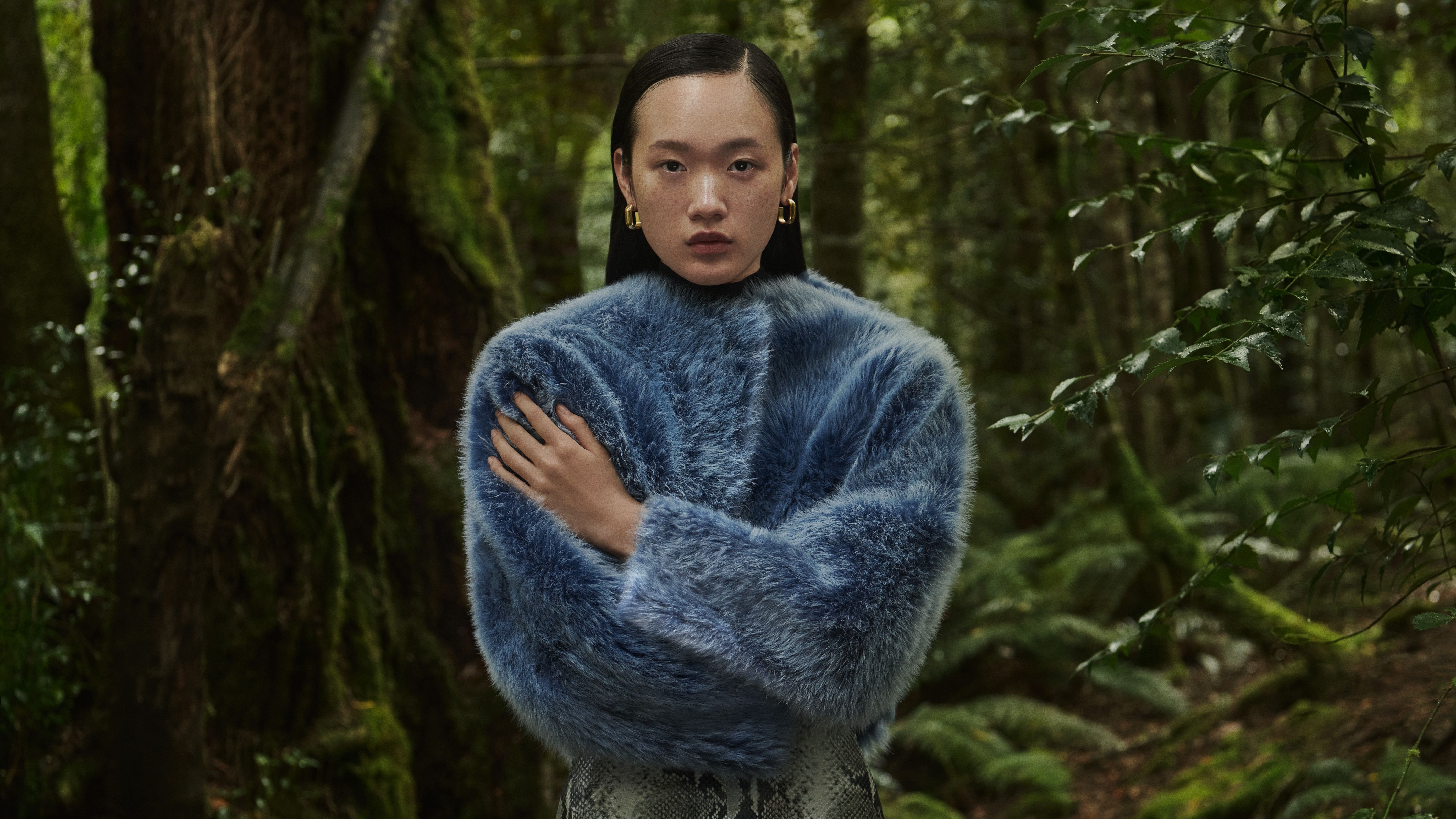 Mid shot of Venus He standing in the forest with arms crossed wearing a blue Toscana shearling jacket