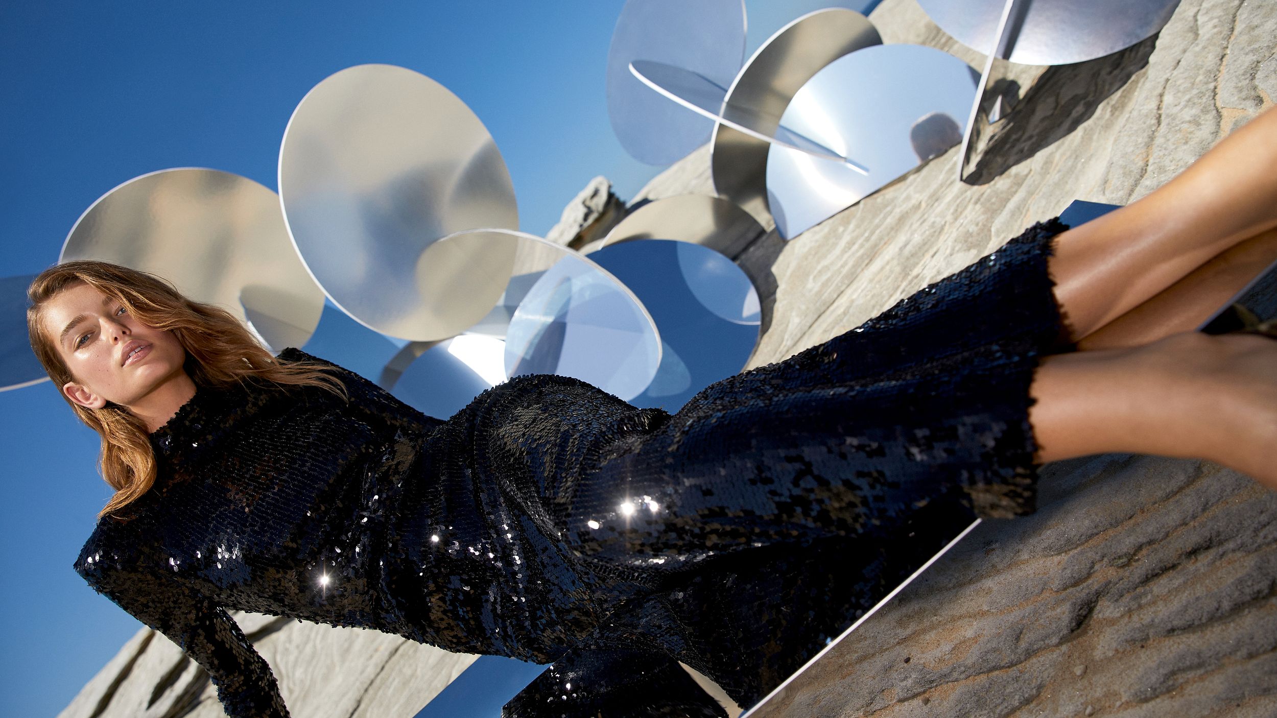 Model reclined amongst mirrored sculptures wearing a black long-sleeved sequin gown