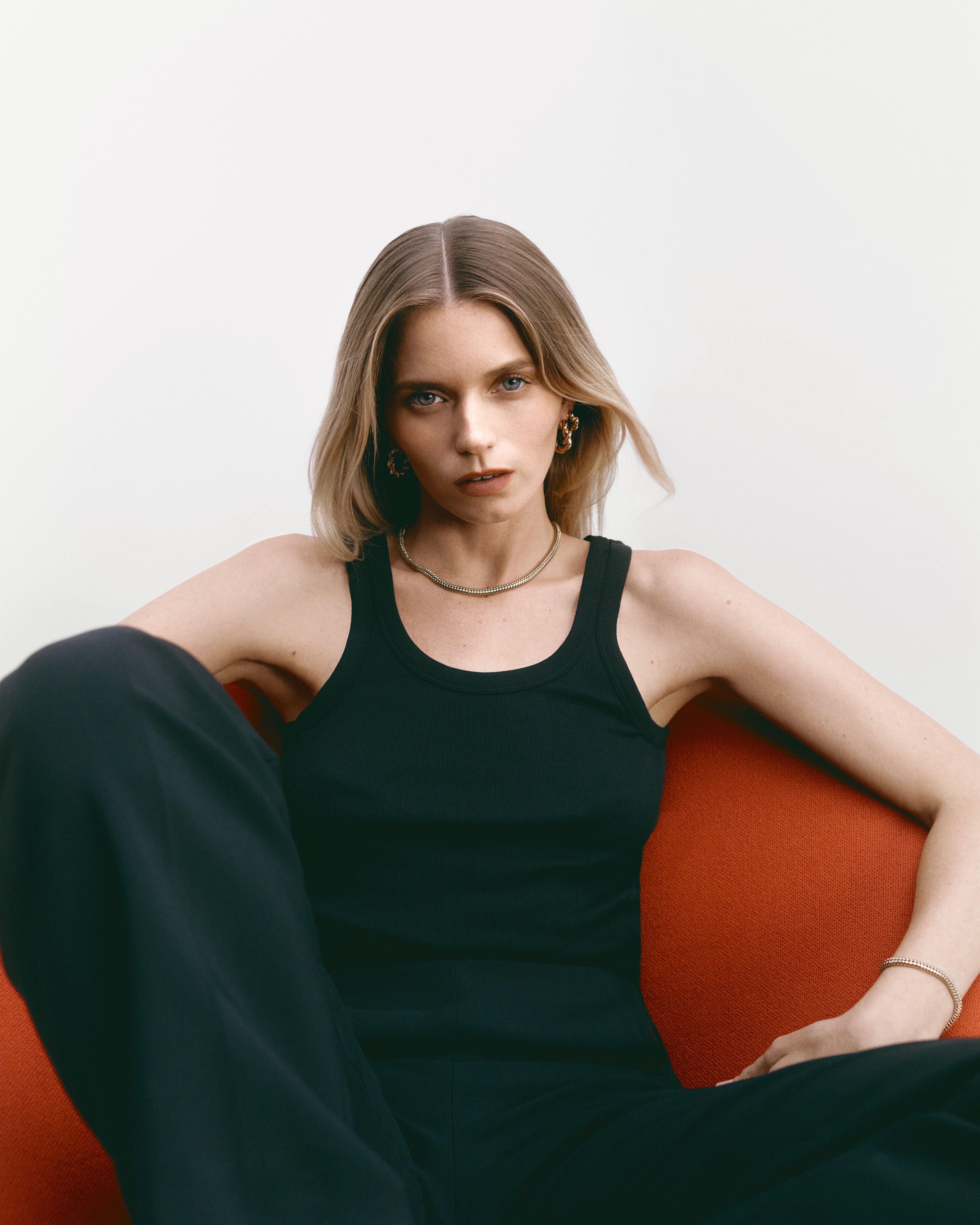 Abbey Lee sitting in a red chair wearing a black tank top and trousers