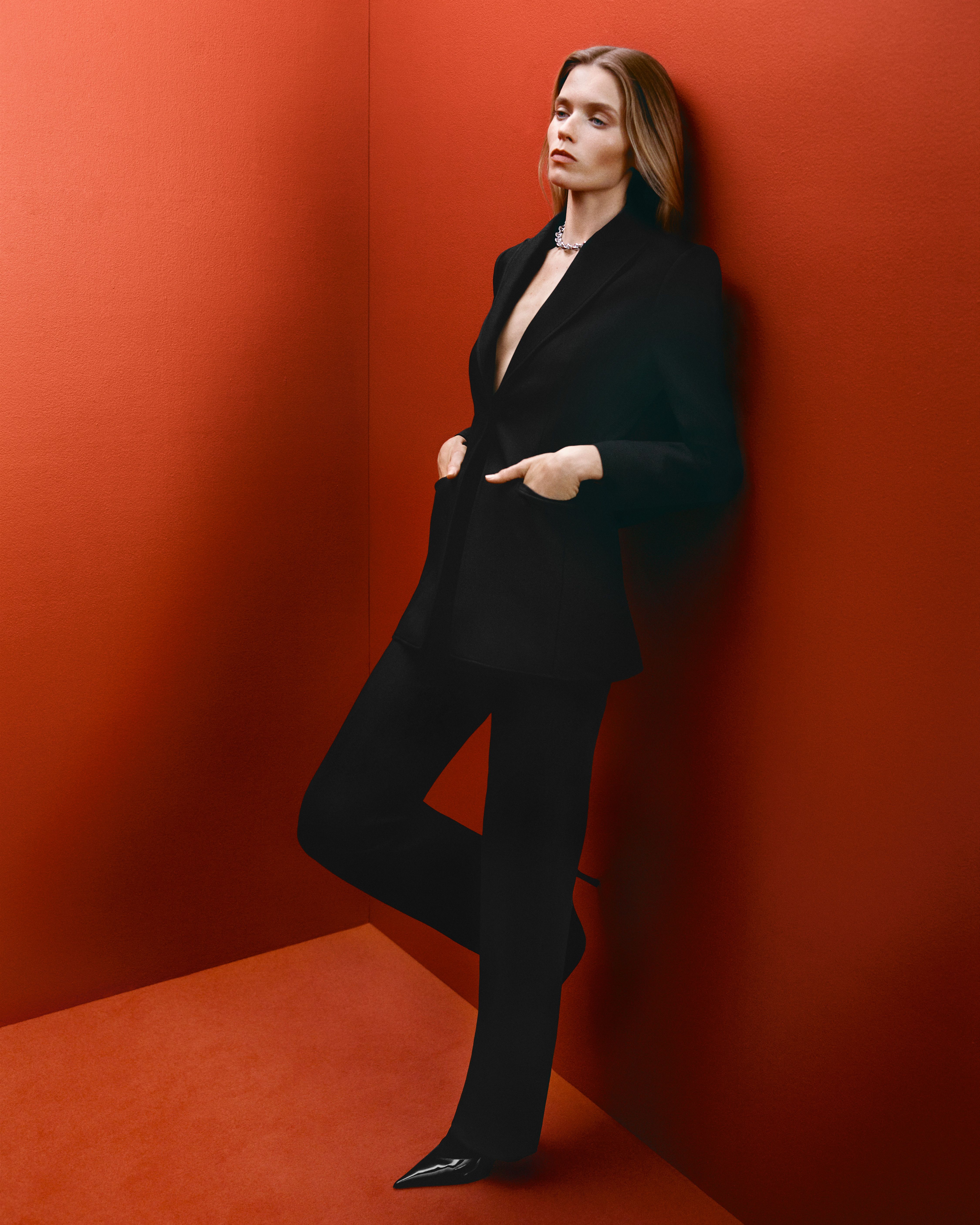 Abbey Lee leaning against the wall in a red room with hands in pockets wearing a black suit