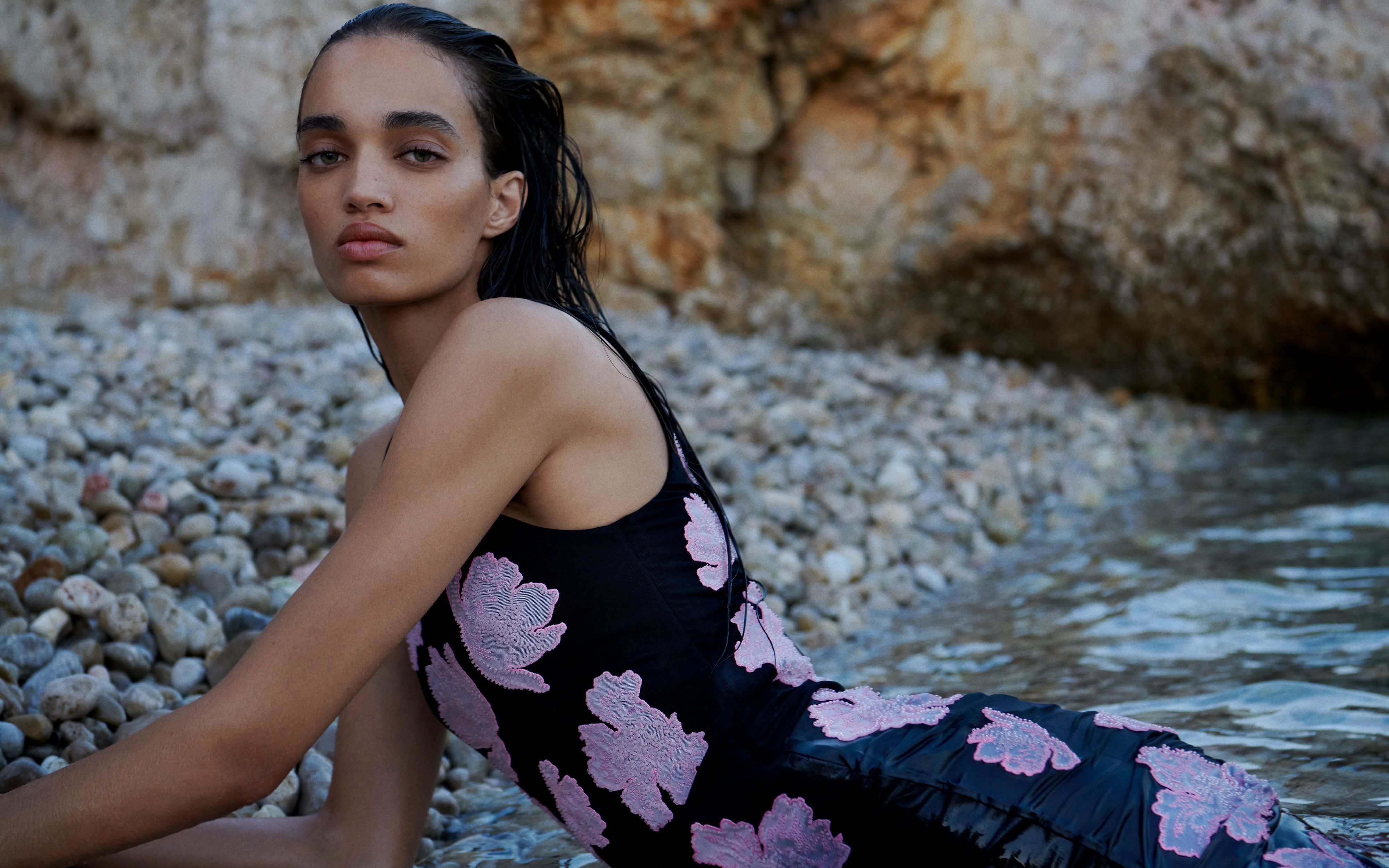 Model reclined on the beach wearing a black dress with embroidered pink flowers