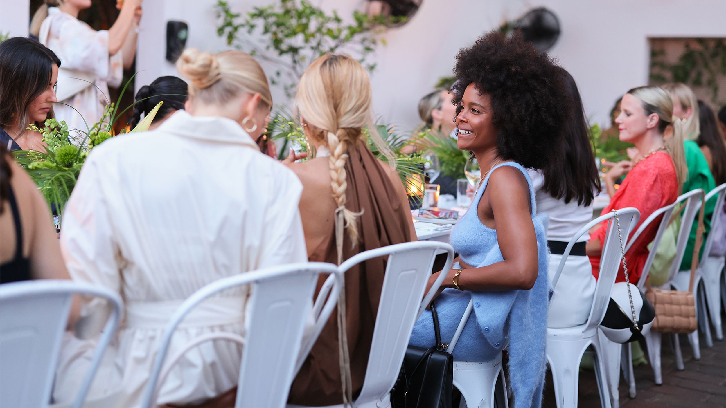 Group of women sitting on white chairs, greenery on table, woman smiling in blue top and skirt