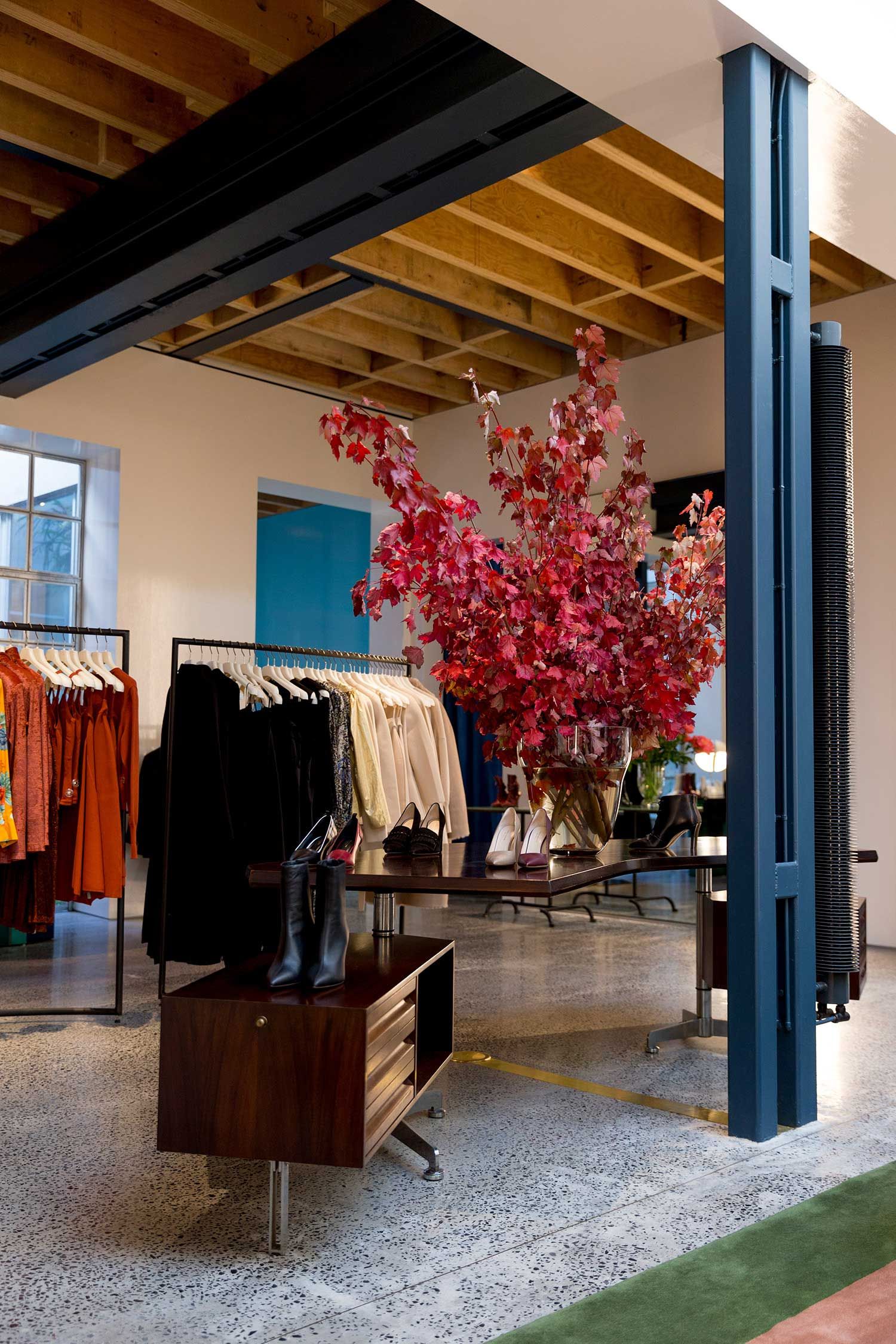 Store display with red leaves floral arrangement, shoes on a desk, and racks of clothing