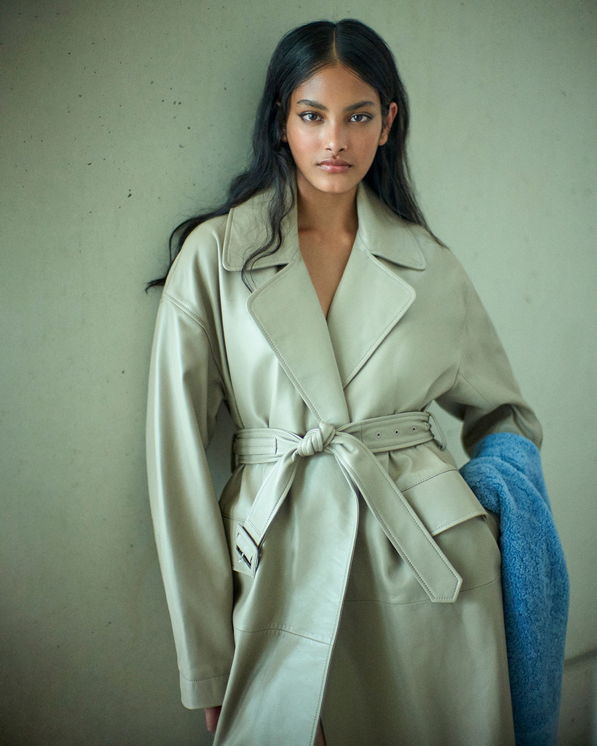 Brunette model leaning against the wall wearing a neutral leather trench and blue scarf