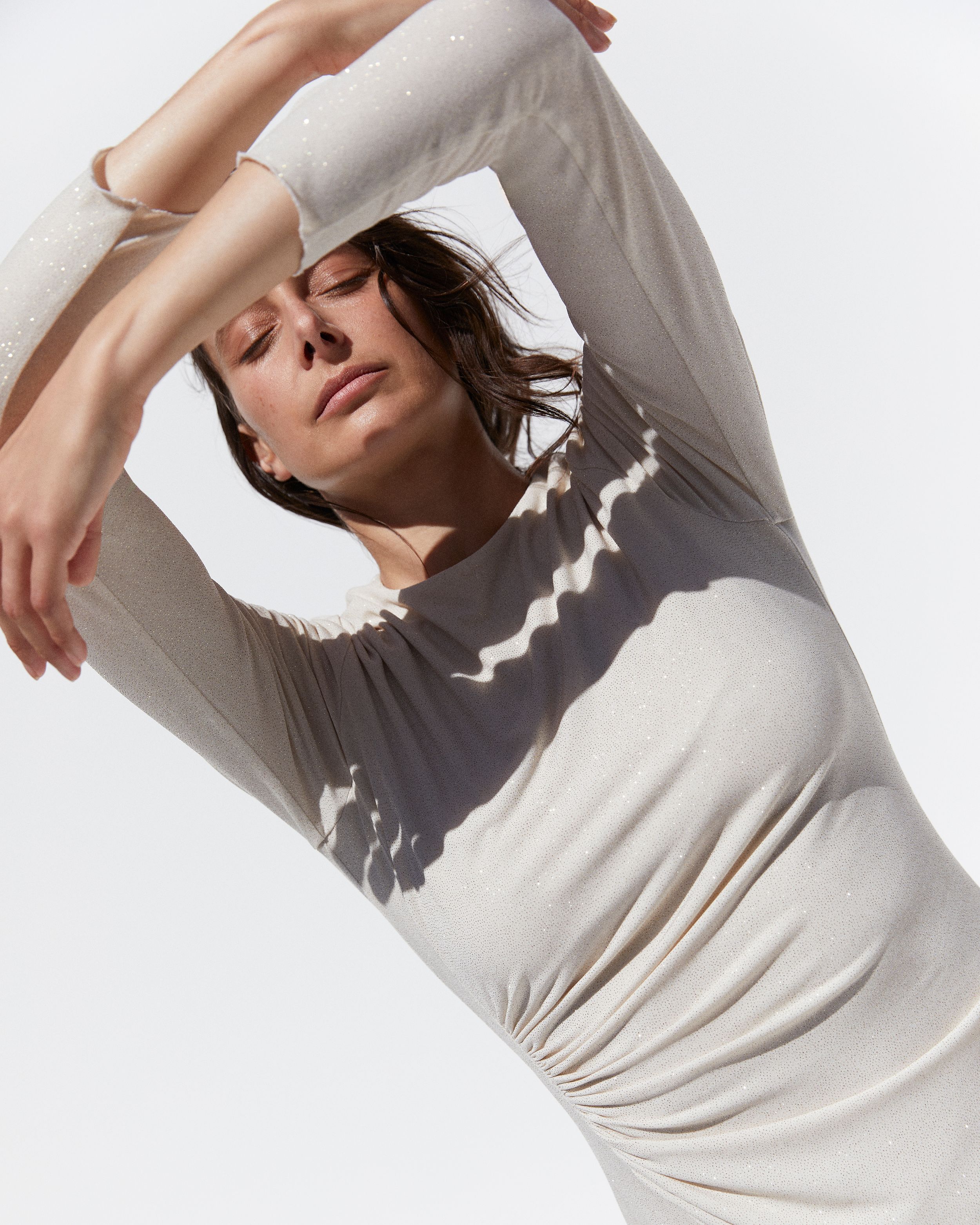 Dimity Azoury posing with eyes closed and arms folded above her head wearing a long-sleeve cream dress