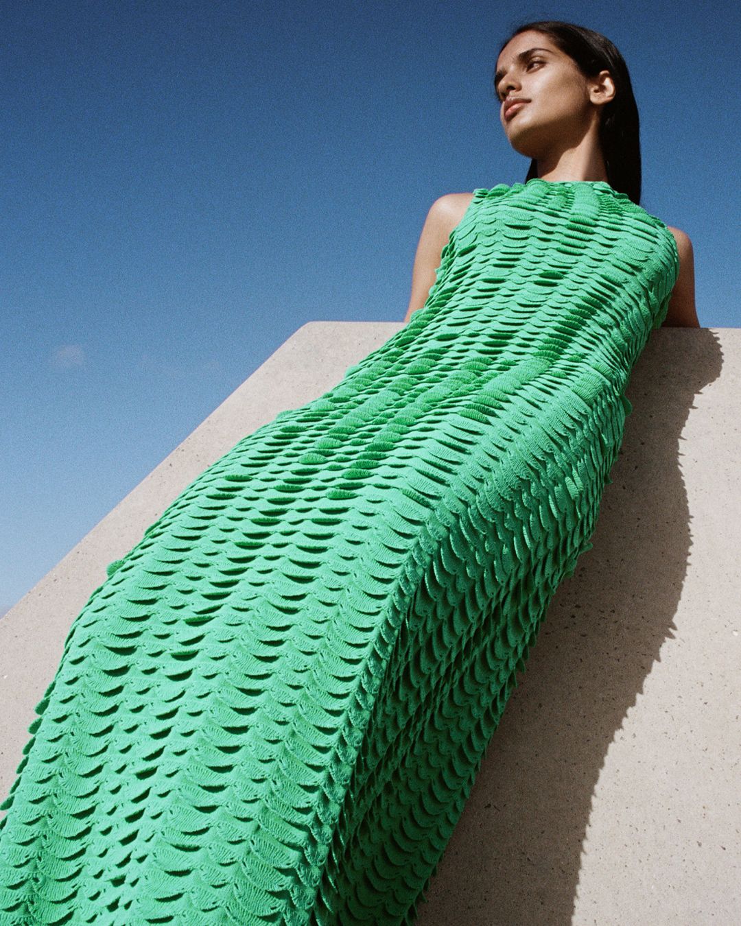 Girl looking away from the camera, leaning against concrete wall in textured green knit dress. 