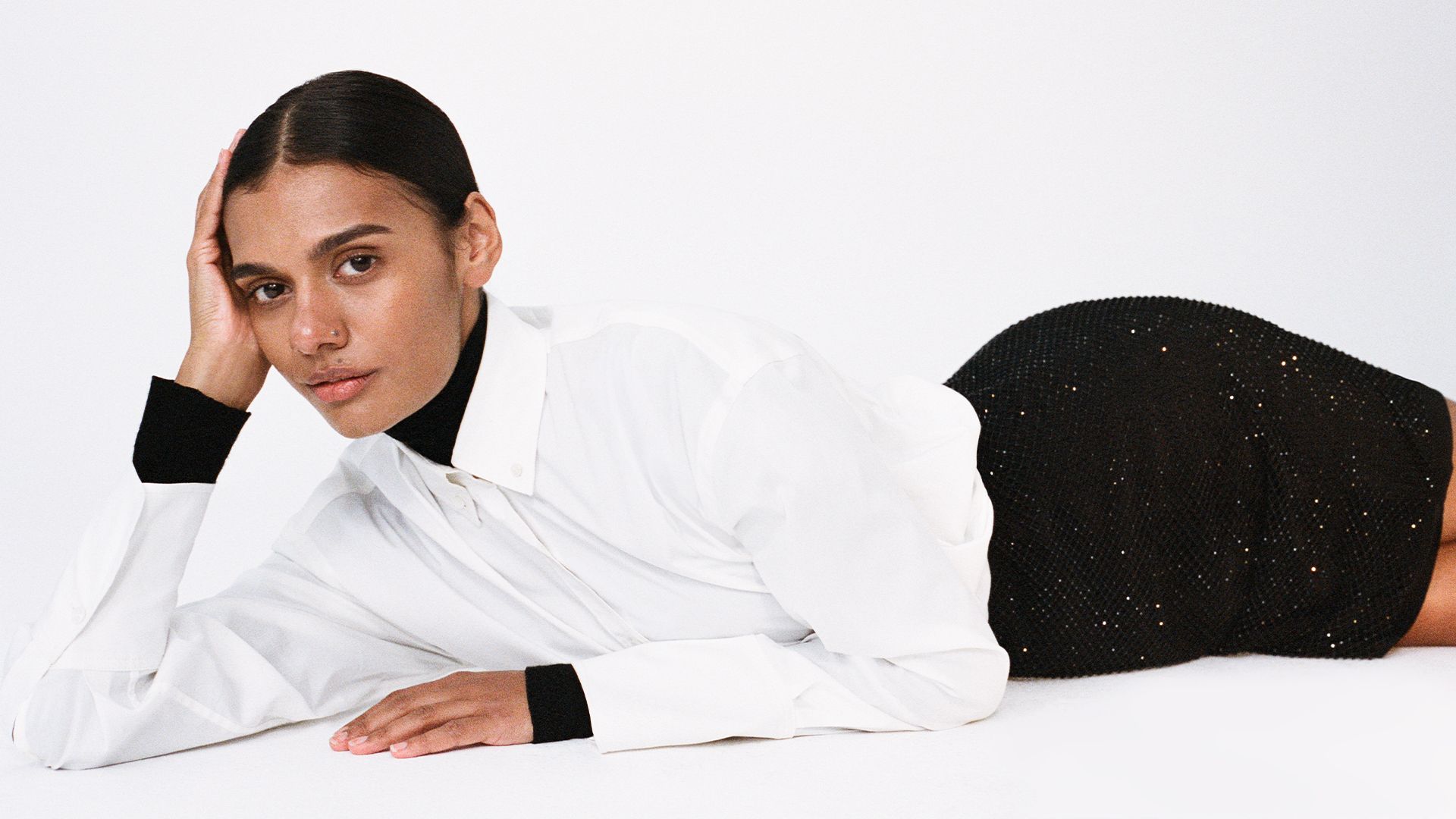 Woman lying down wearing a white cotton shirt, layered with a black babywool turtleneck and sparkly black mini skirt