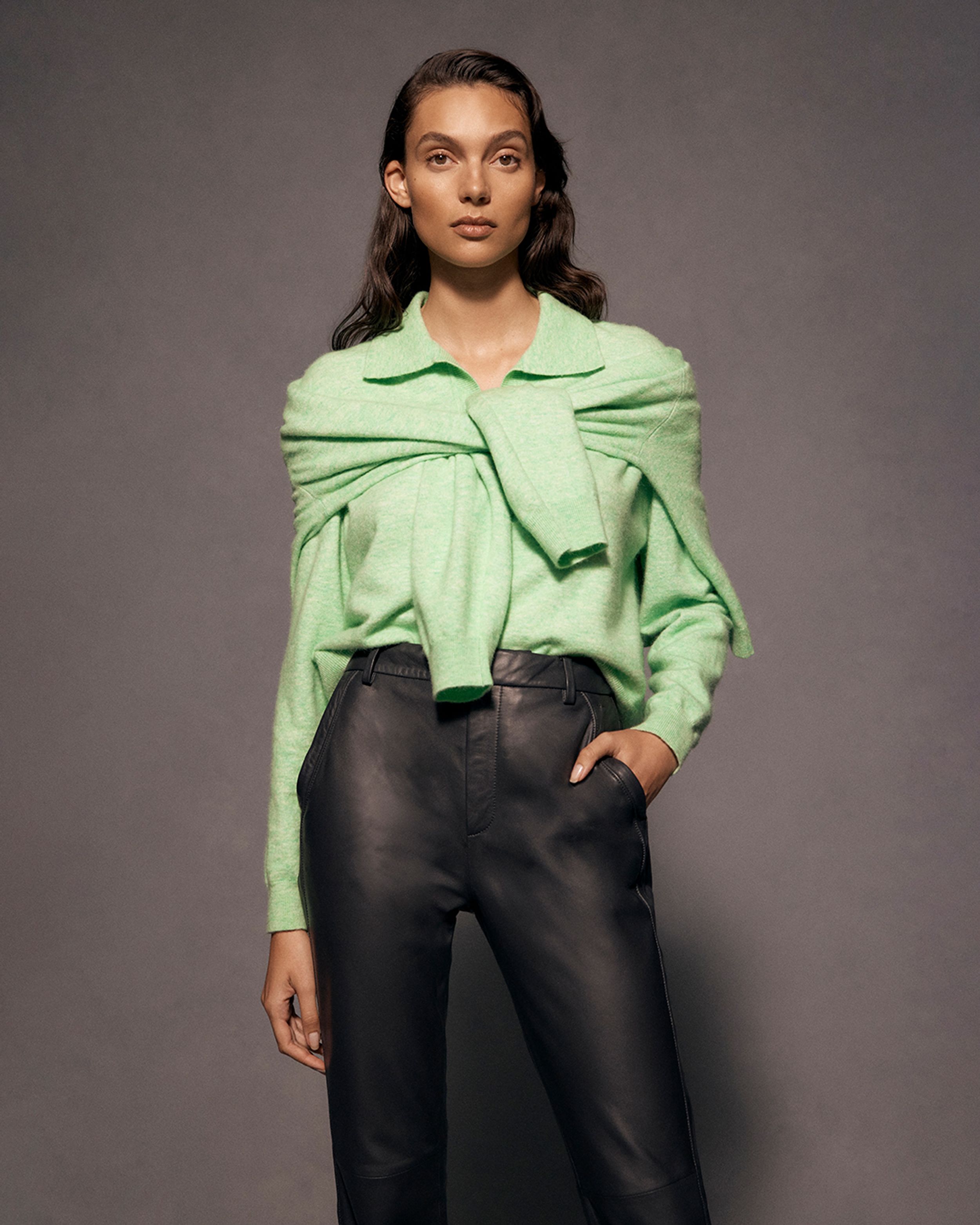 Brunette model posing with hand in pocket wearing a green knit and black trousers